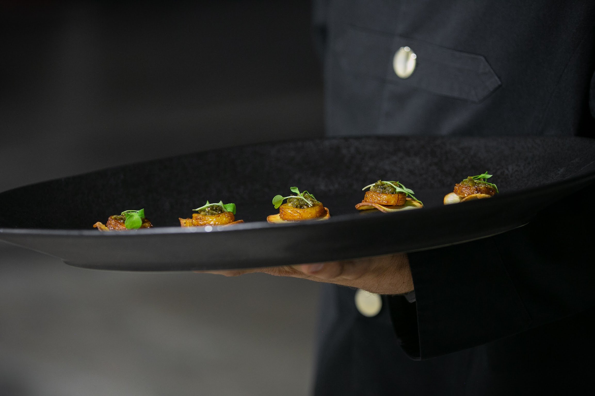 A tray with five small appetizers or finger foods, each topped with greens, being held by a person in a black shirt with a visible pocket and button.