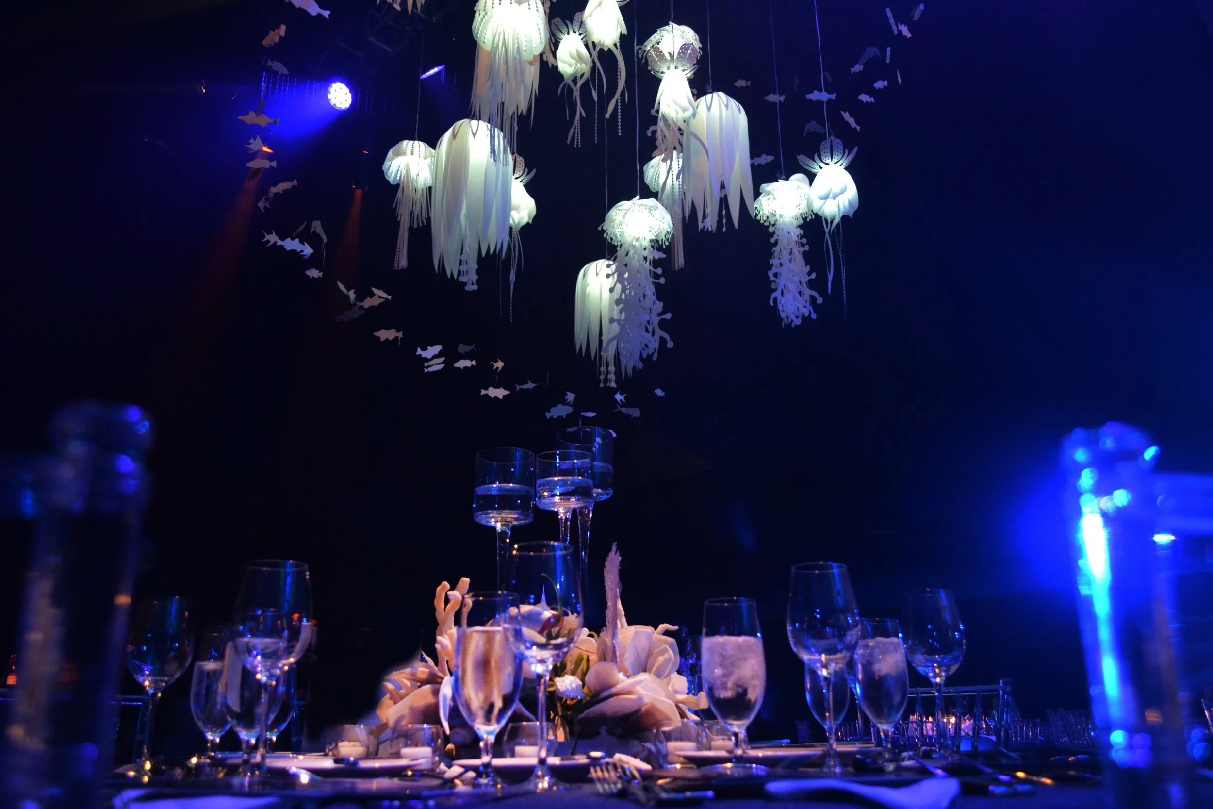 Elegantly set table with wine glasses, floral centerpiece, and hanging jellyfish-shaped lights in a dark, blue-lit environment.