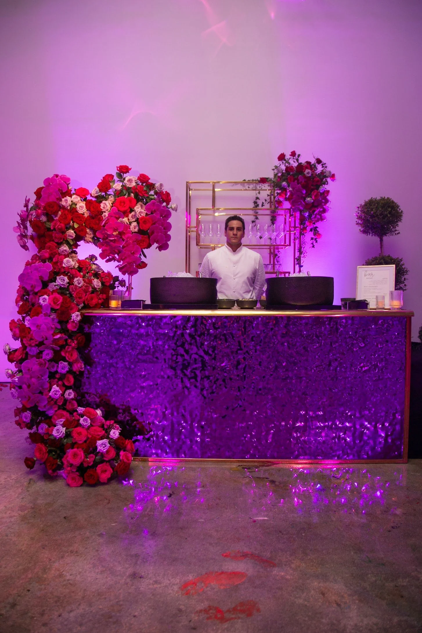 A bar decorated with a large bouquet of pink and red flowers, with purple lighting and a chef standing behind the counter.