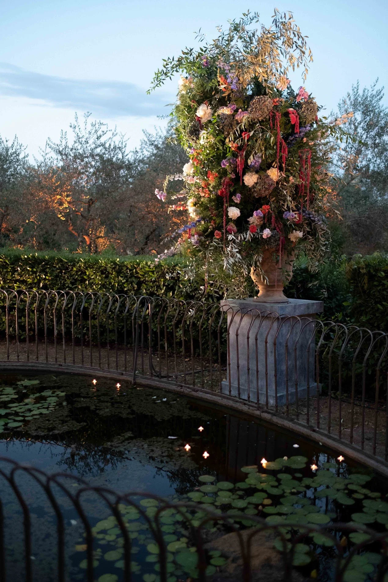 A large decorative floral arrangement in a tall stone vase on a pedestal, situated outdoors near a pond with floating candles and lily pads. Mature trees and a cloudy sky are in the background.