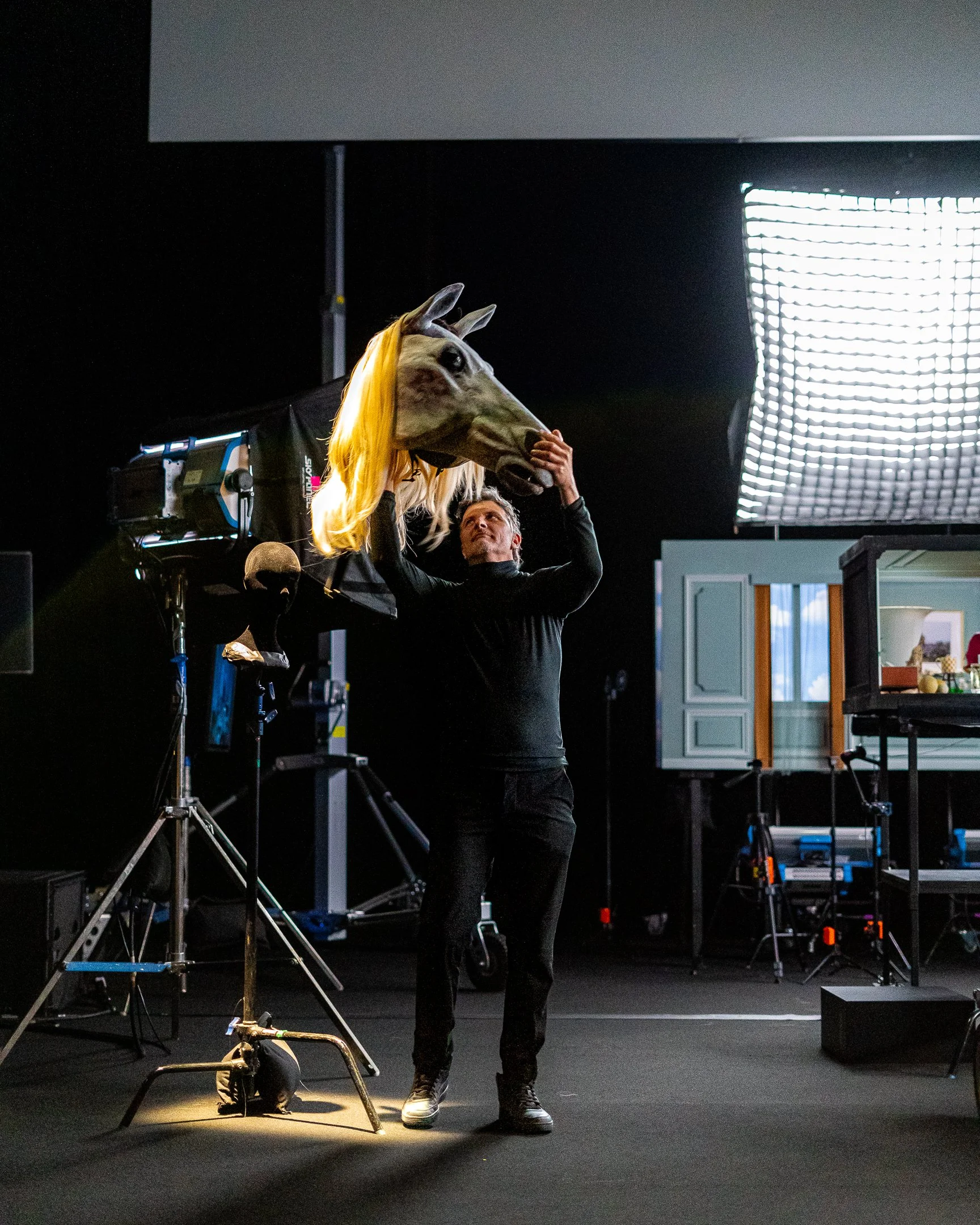 A person wearing a horse mask is holding a video camera on a tripod in a studio setting, with stage lighting equipment and a kitchen area visible in the background.