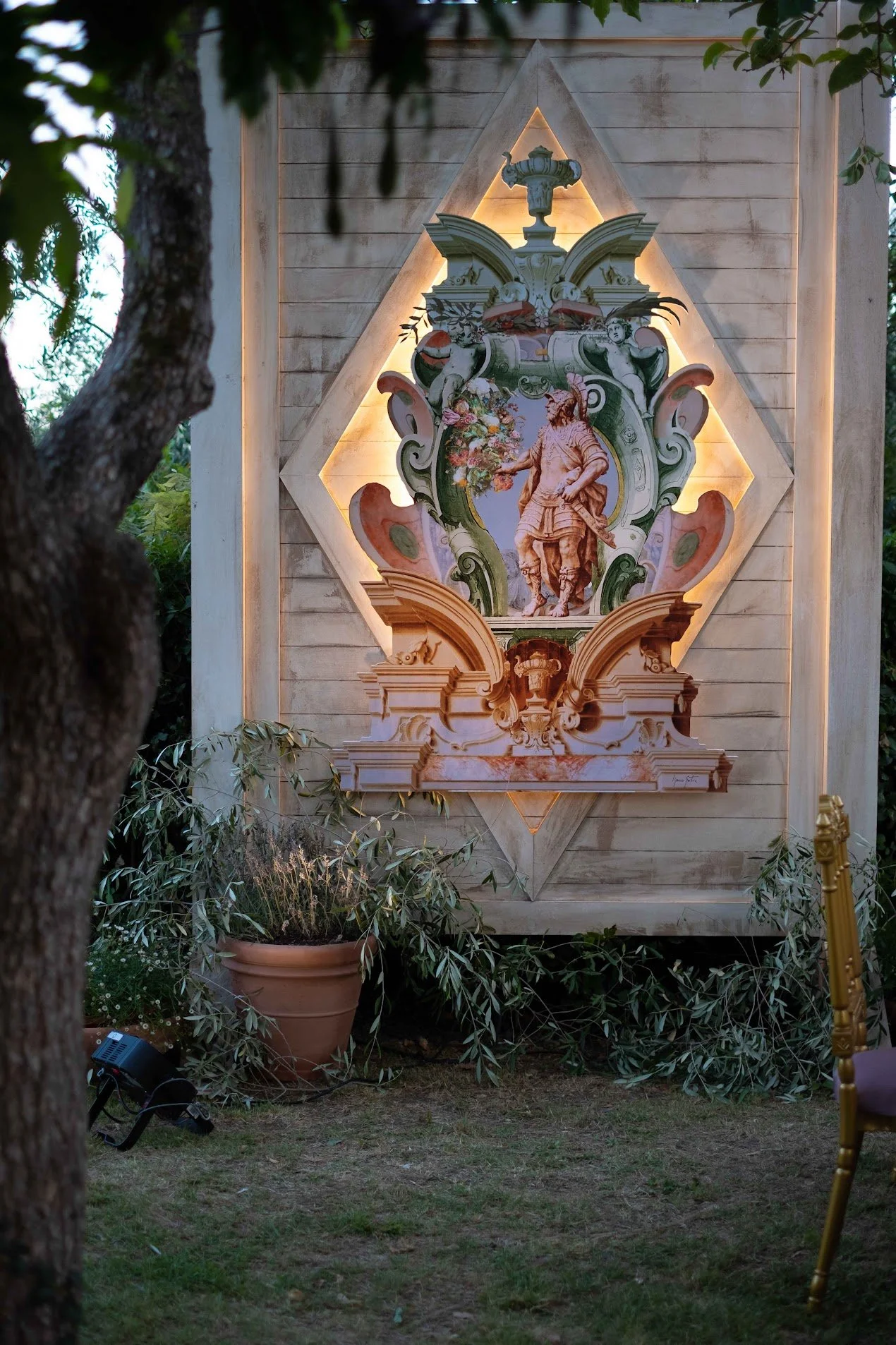 A decorative religious or heraldic mural with a central figure of a man holding flowers, framed by ornate scrollwork and cherubs, illuminated against a wooden wall, surrounded by potted plants and trees.