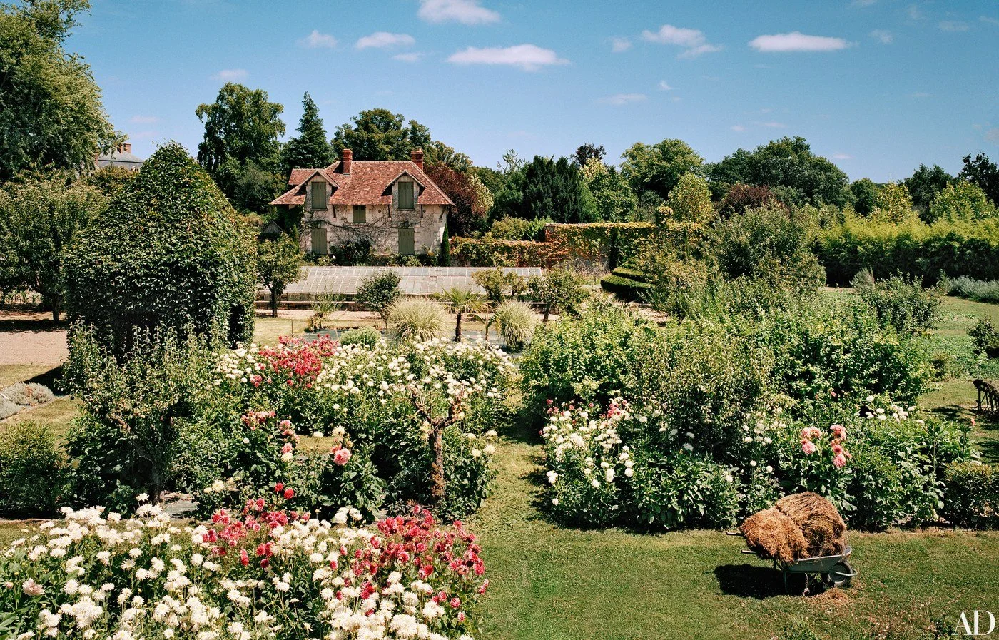 A lush garden venue for special events in France with blooming flowers and trees, a small wheelbarrow loaded with hay, and a stone house with a red-tiled roof in the background.