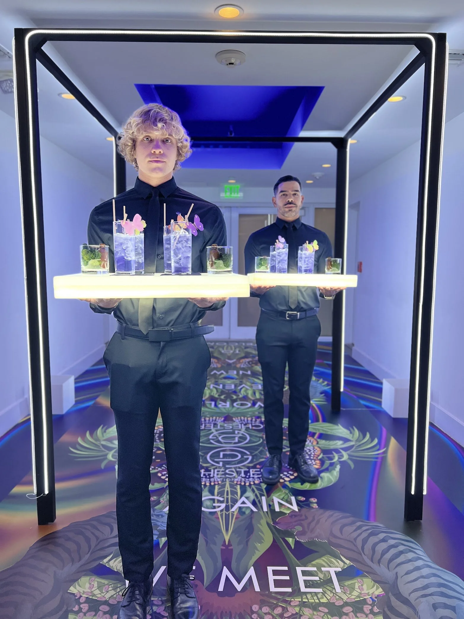 Two men dressed in black shirts and black pants holding trays with drinks, standing in front of a decorative frame with neon lighting inside a lobby or event space.