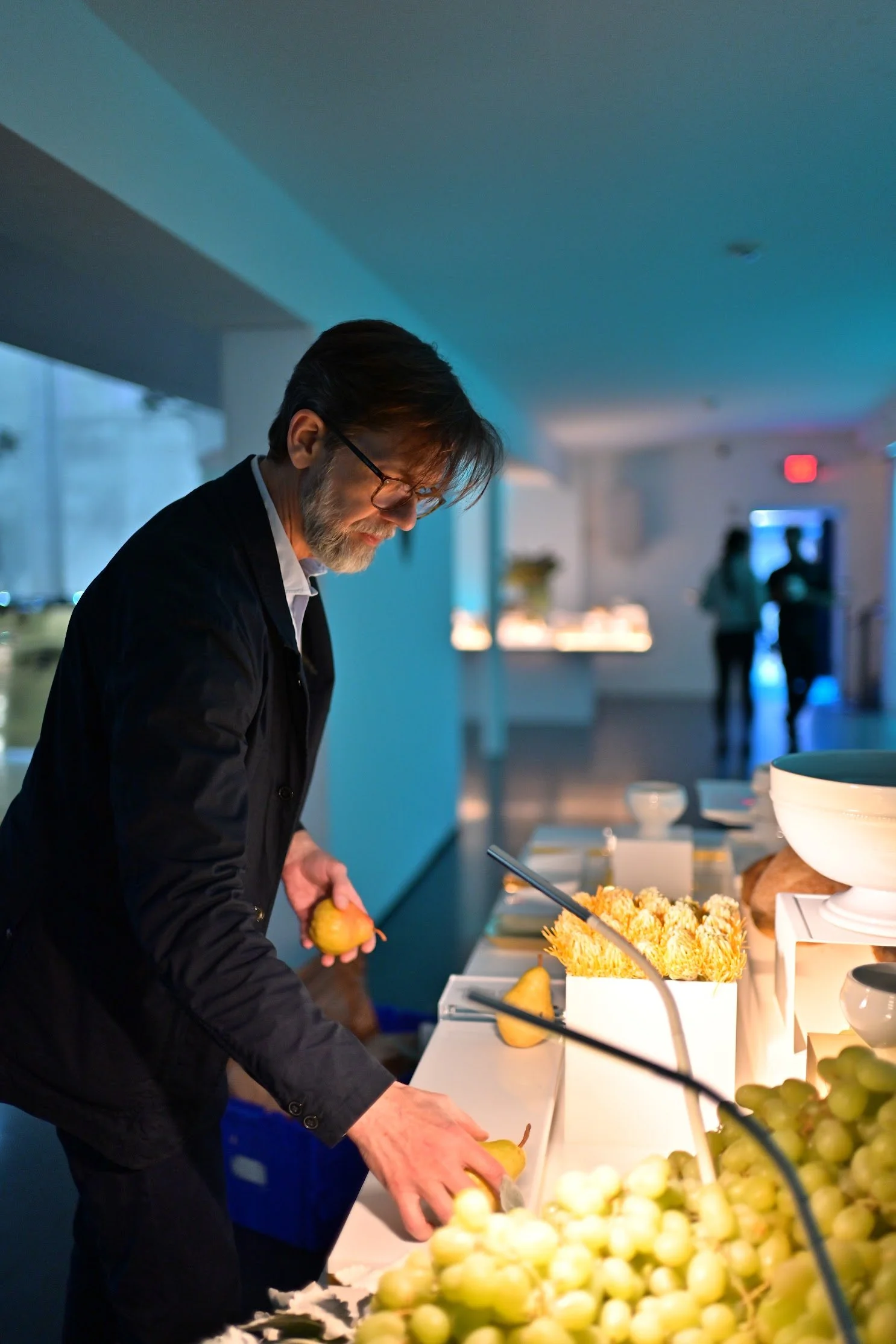 Man with glasses and beard selecting fruit at a buffet table with grapes and other items, in a modern indoor setting.