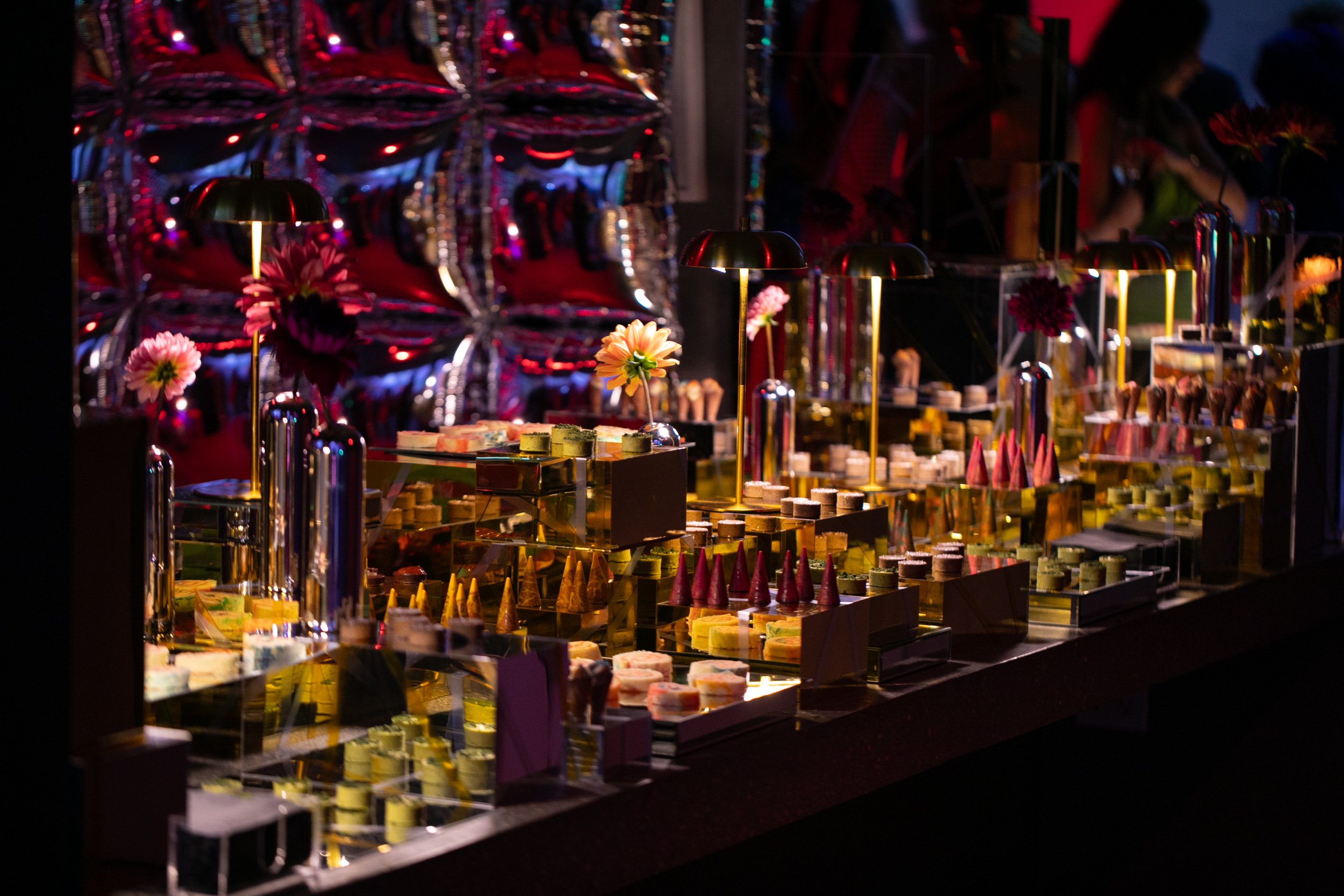 Colorful candy display with various sweets, flowers, and illuminated metal lamps at a checkout counter in a festive or party setting.