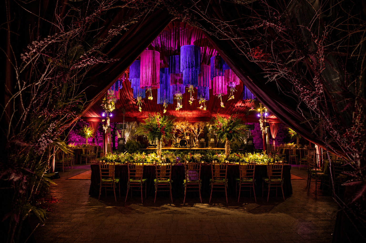 Decorated event venue with vibrant purple and pink hanging lanterns, tropical plants, and a stage set for a celebration.