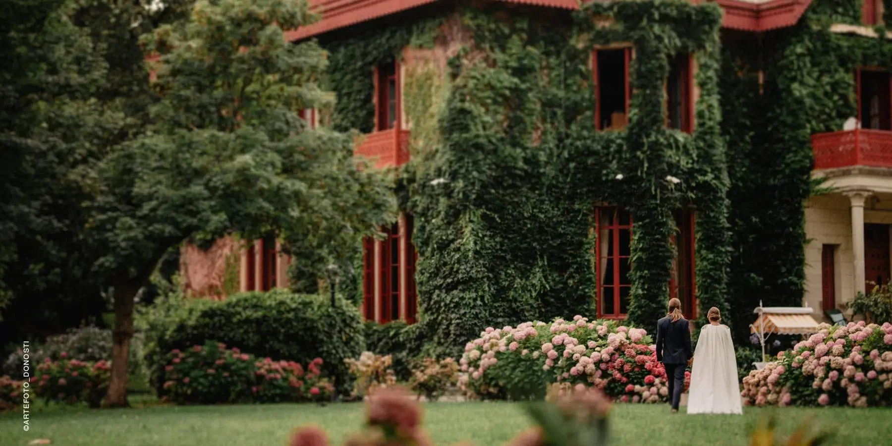 A couple walks in a garden at the Palacio de Arbaisenea, with pink hydrangeas in front of a building covered in green ivy, with red-trimmed windows and balconies.