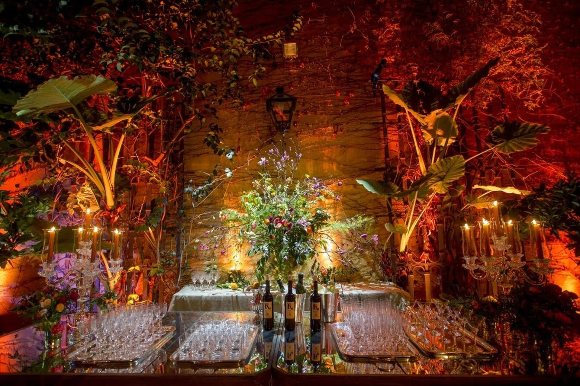Table setting with glasses, bottles, and flowers at an event with greenery, candles, and warm lighting on a decorated wall.
