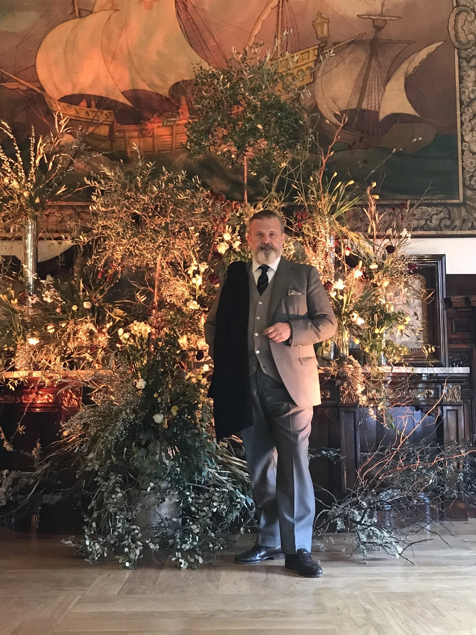 A man with a beard and mustache dressed in a gray suit and tie, standing in front of a large floral arrangement with greenery and flowers, and a backdrop of a mural depicting old sailing ships.