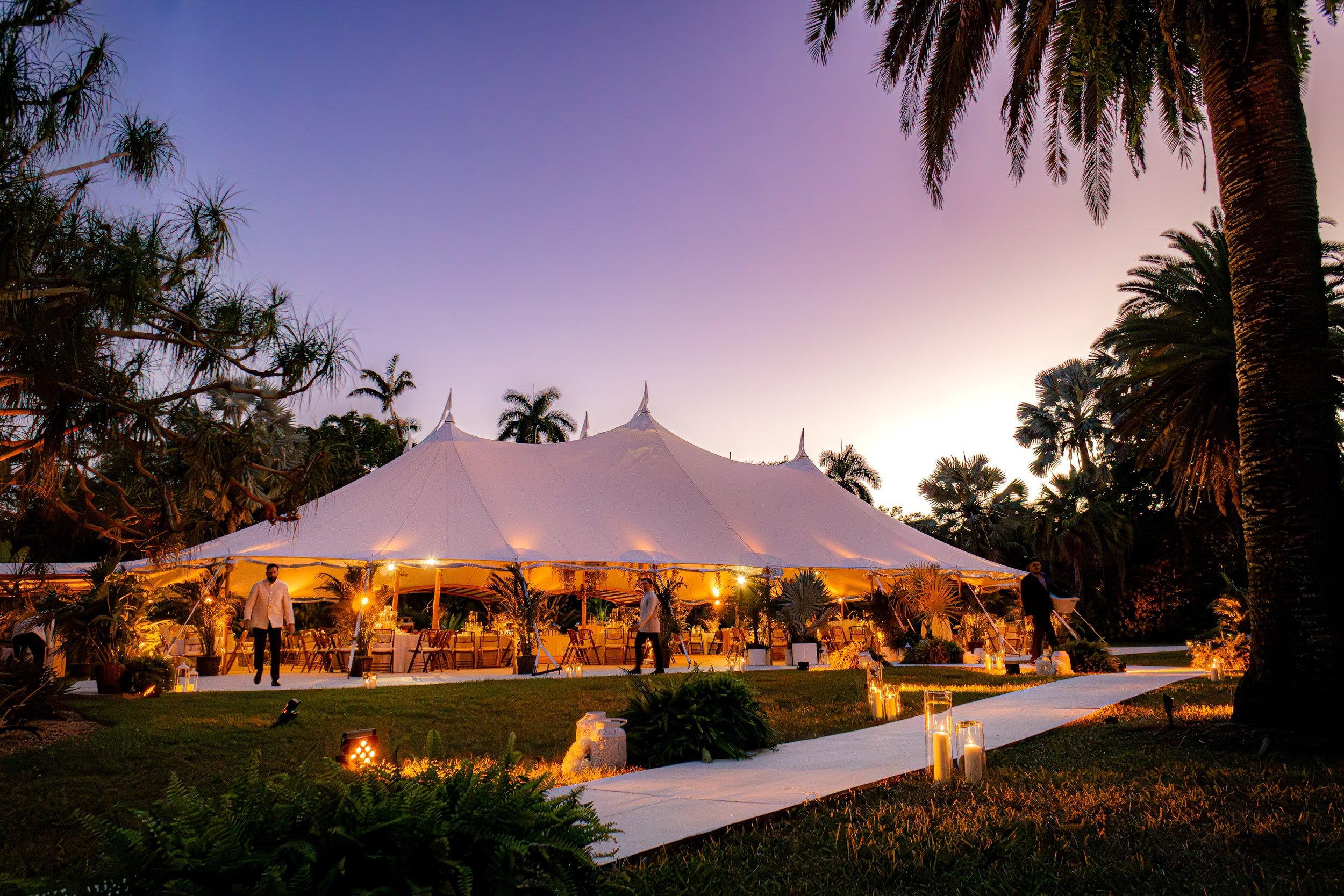 An outdoor event under a white tent with a purple evening sky, surrounded by tall palm trees and lush greenery, lit by warm string lights and candles.