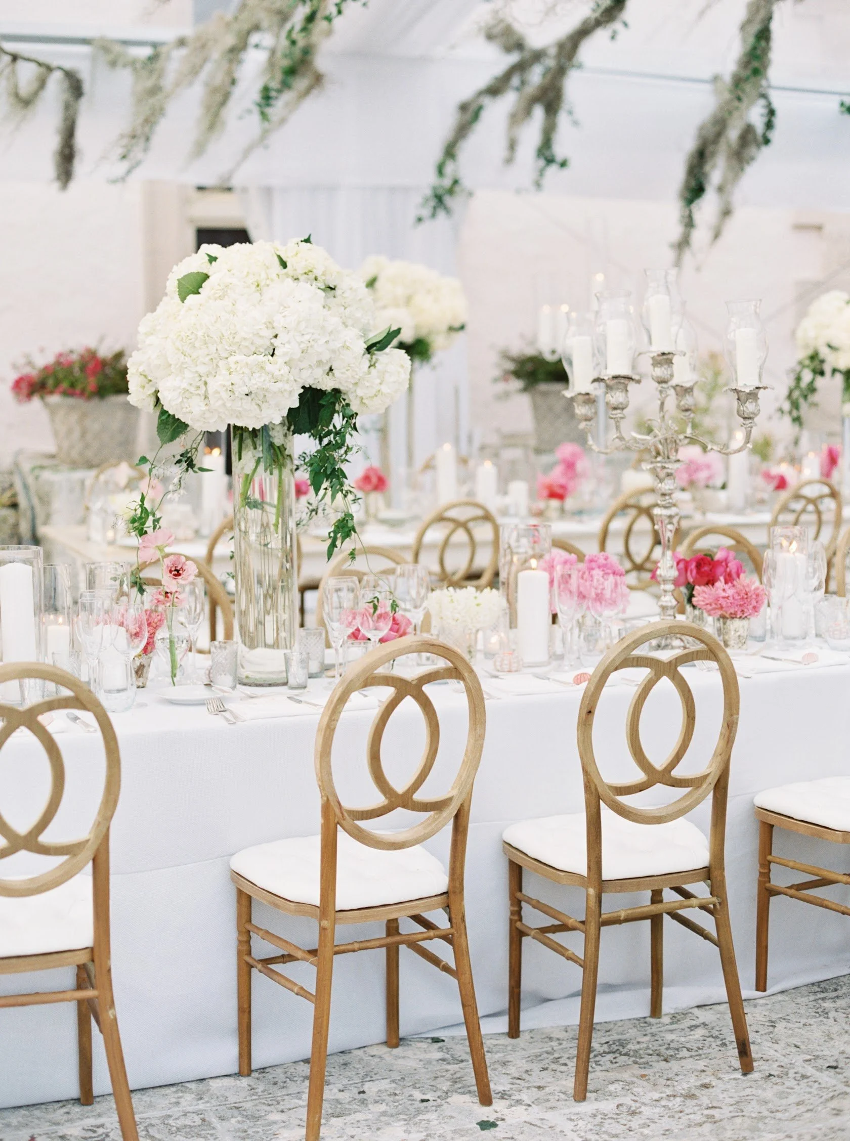 Wedding reception table decorated with white and pink flowers, candles, and a large floral centerpiece. Wooden chairs surround the table, with hanging greenery above.