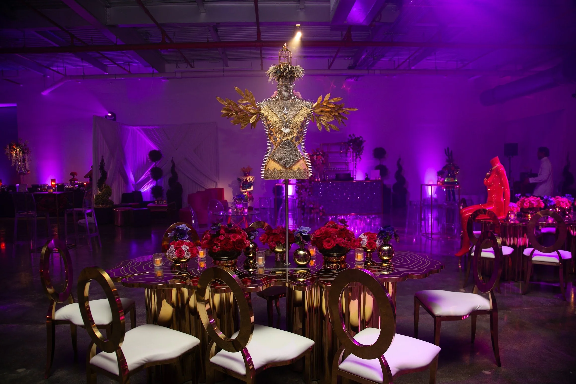 An event space decorated with a large chandelier adorned with golden and silver accents, sitting in the center surrounded by pink and red floral arrangements. The room is illuminated with purple and pink lighting, and tables with chairs are arranged around the chandelier.