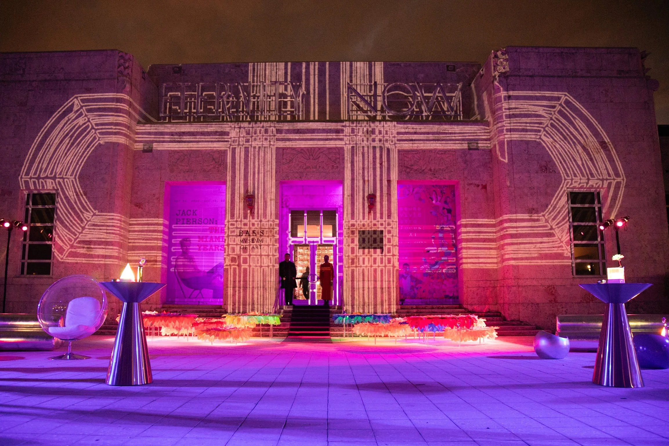 Nighttime view of a building facade with pink and purple lighting, projection of abstract lines and text, and a few people standing at the entrance, with tables and chairs in the foreground.