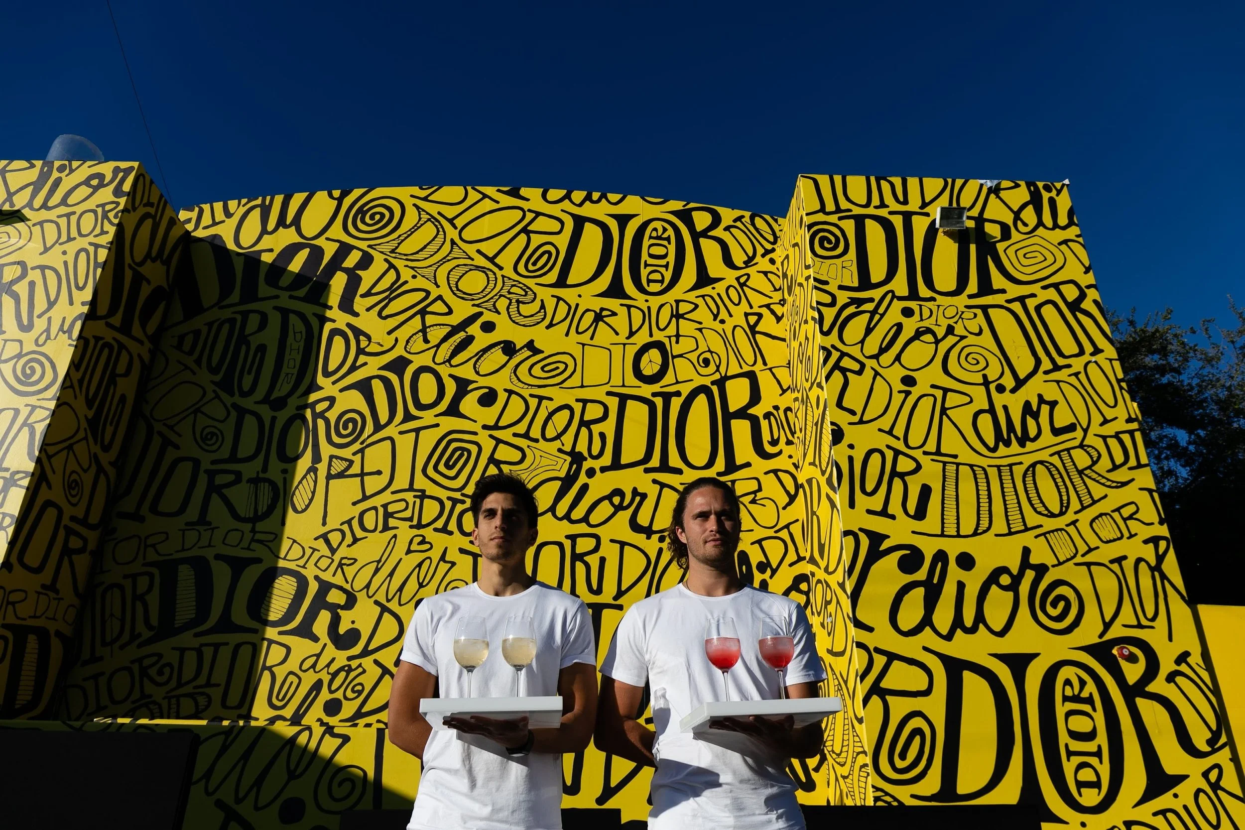 Two men in white shirts holding trays with glasses of wine, standing in front of a bright yellow wall covered in black graffiti text reading 'Dior' repeatedly, with a clear blue sky overhead.