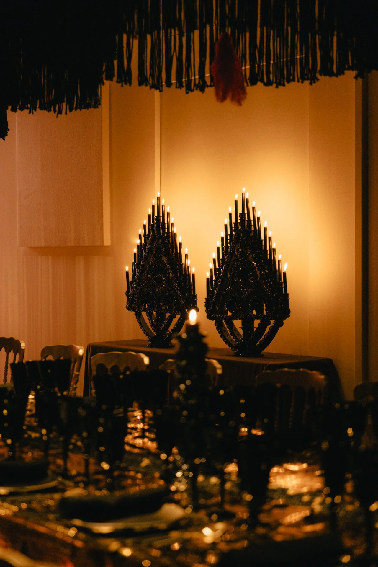 Dimly lit dining room with black candleholders and candles on a table, warm yellow lighting on the wall, decorative tassels hanging from the ceiling, and chairs around the table.