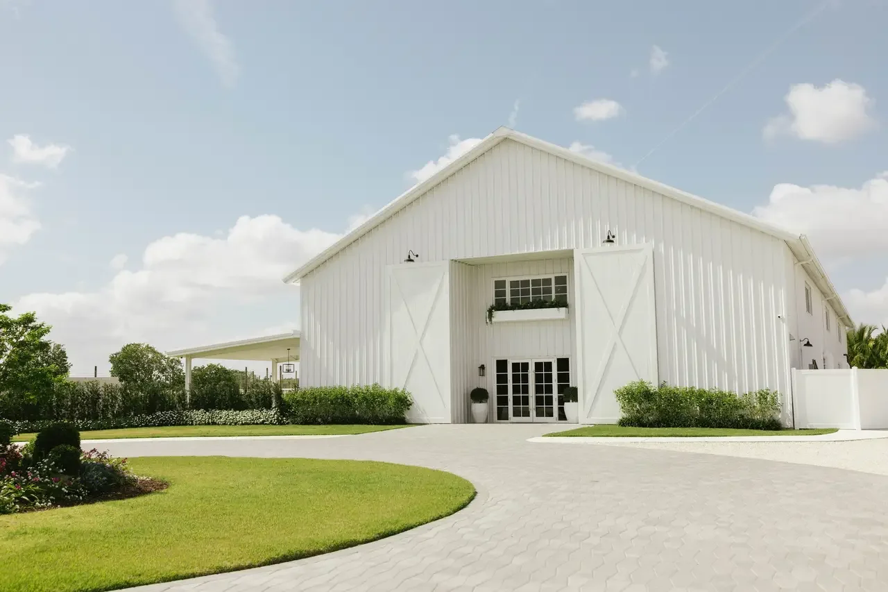 White barn-style building with large double doors, surrounded by green lawns and shrubs, under a partly cloudy sky.