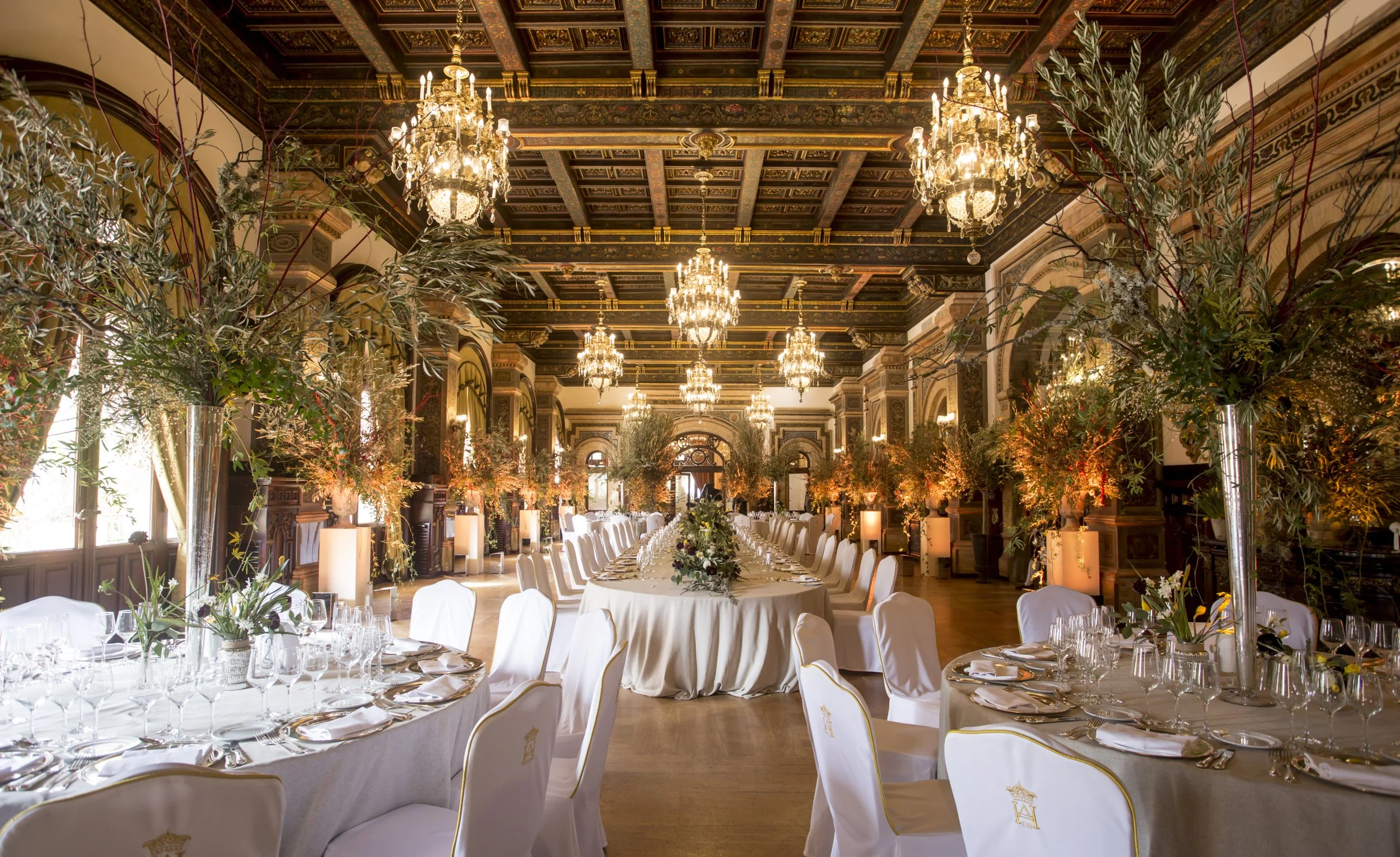 An elegant banquet hall with lavish chandeliers hanging from a decorative ceiling, decorated for a formal event with long tables covered in white linens, arranged with glassware, silverware, and floral centerpieces, surrounded by white chairs with gold accents, and lush greenery and floral arrangements.