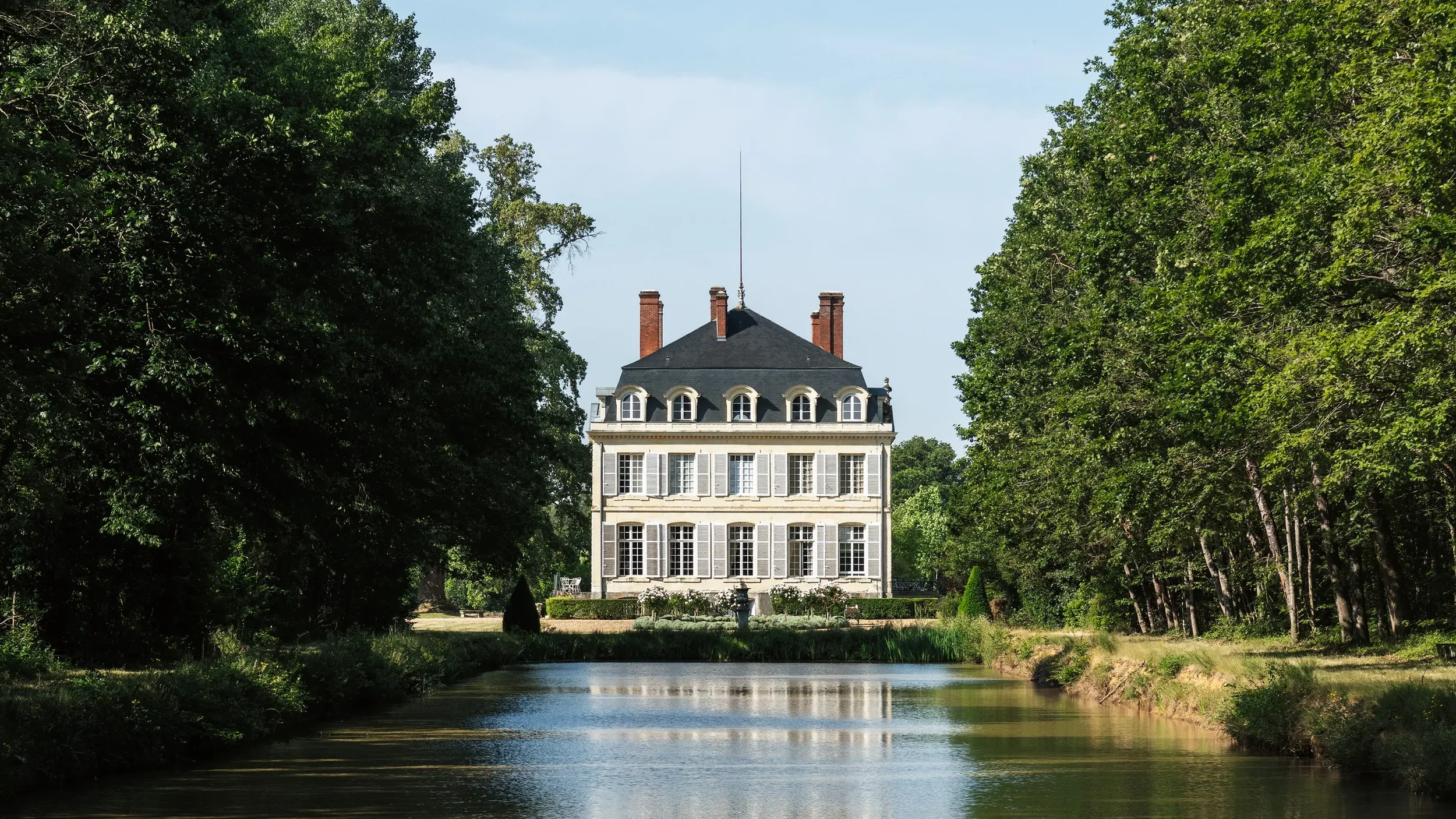 A large, elegant château  with multiple floors and many windows, surrounded by lush green trees and a body of water in the foreground. The location is privately held but can host exclusive luxury events.