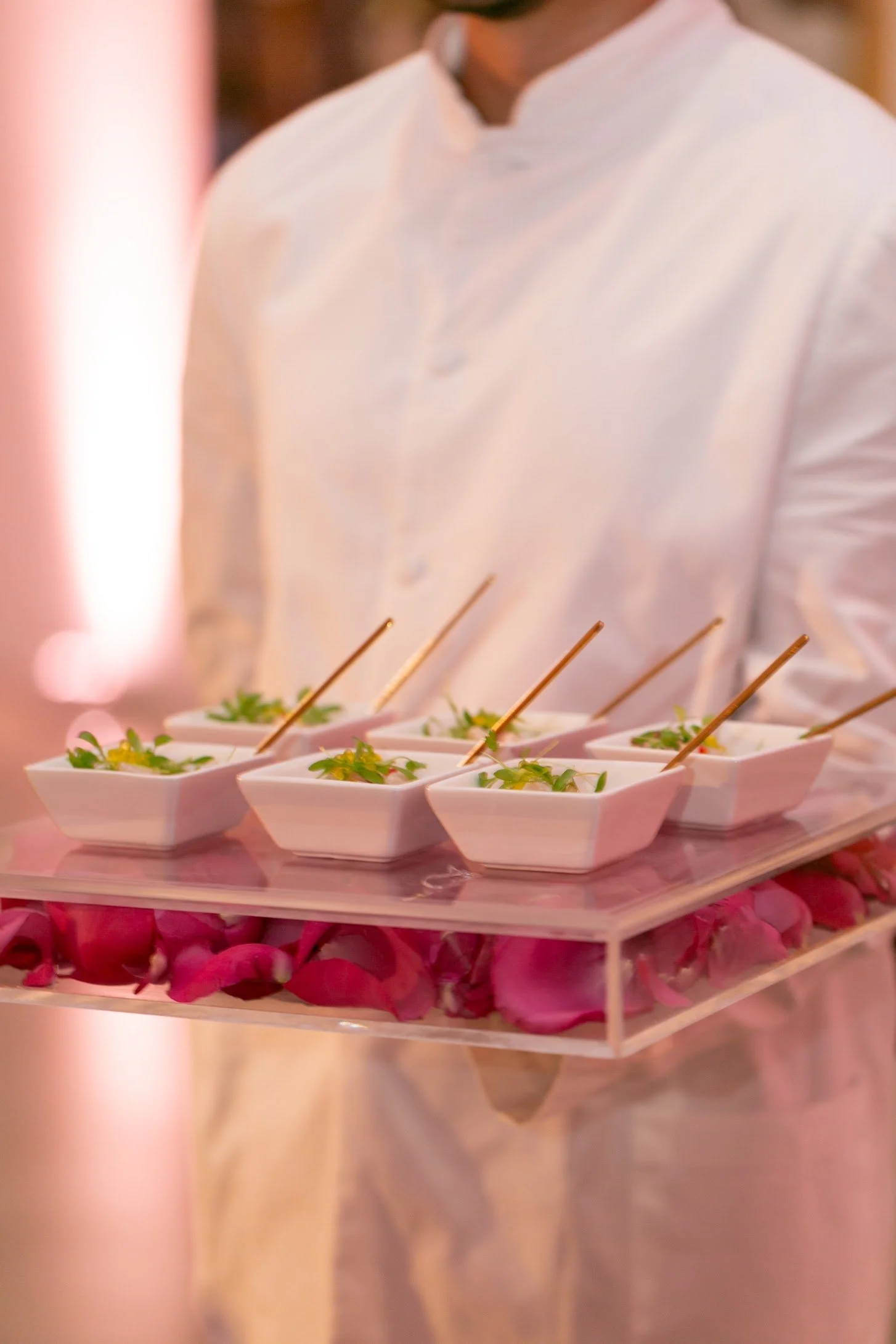 A person wearing a white shirt holding a tray of small white dishes with food garnished with herbs on a bed of pink flower petals.