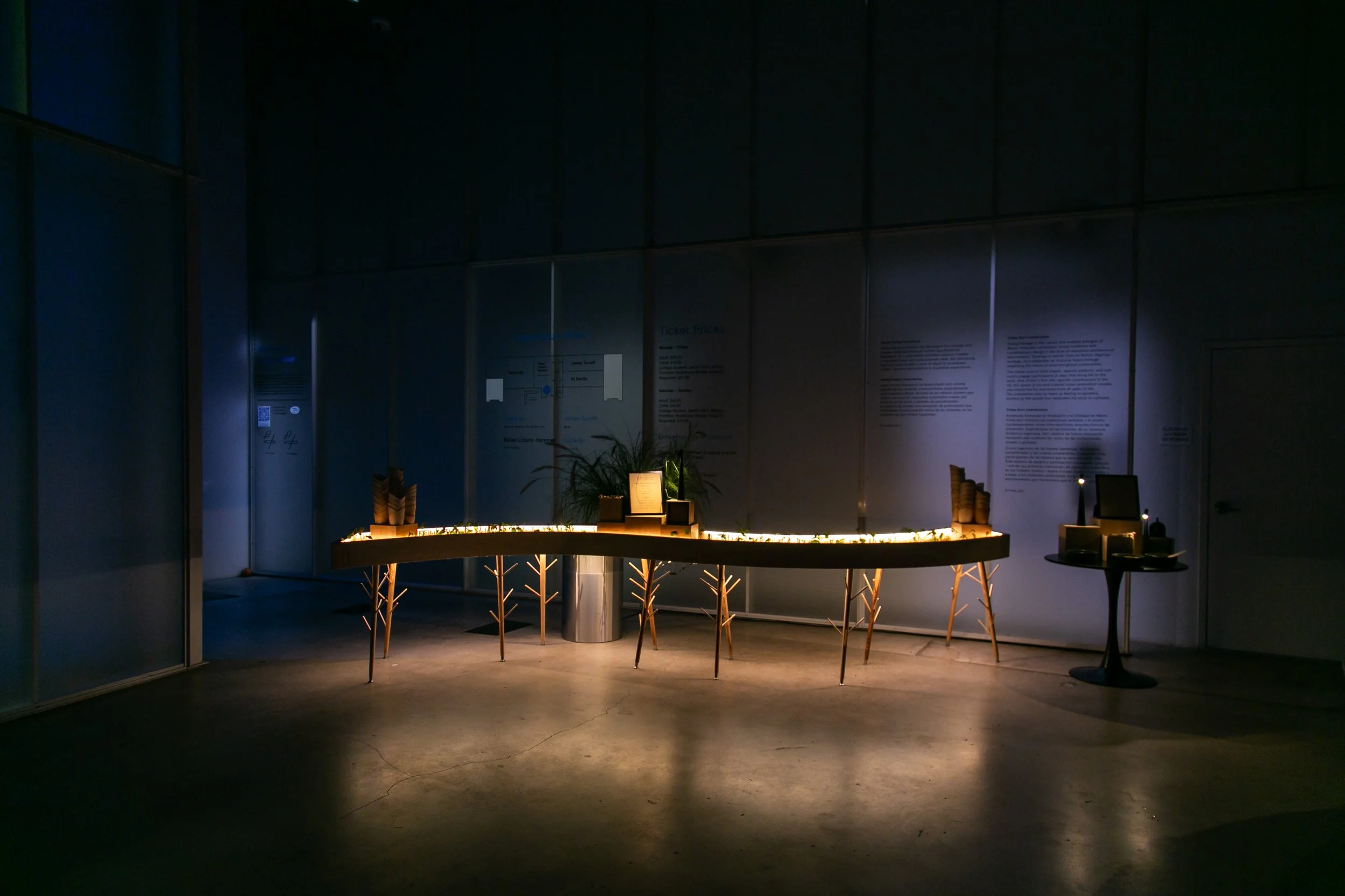 A dimly lit interior space featuring a round wooden table with decorative items on top, surrounded by chairs, and illuminated by a small light. The background has gray walls with informational panels and a tall plant in the center.