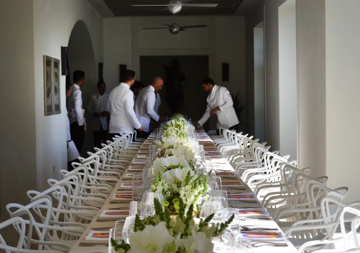Long dining table decorated with white flowers, set with glasses, plates, and cutlery in a bright indoor space, with several people in white attire preparing the table.
