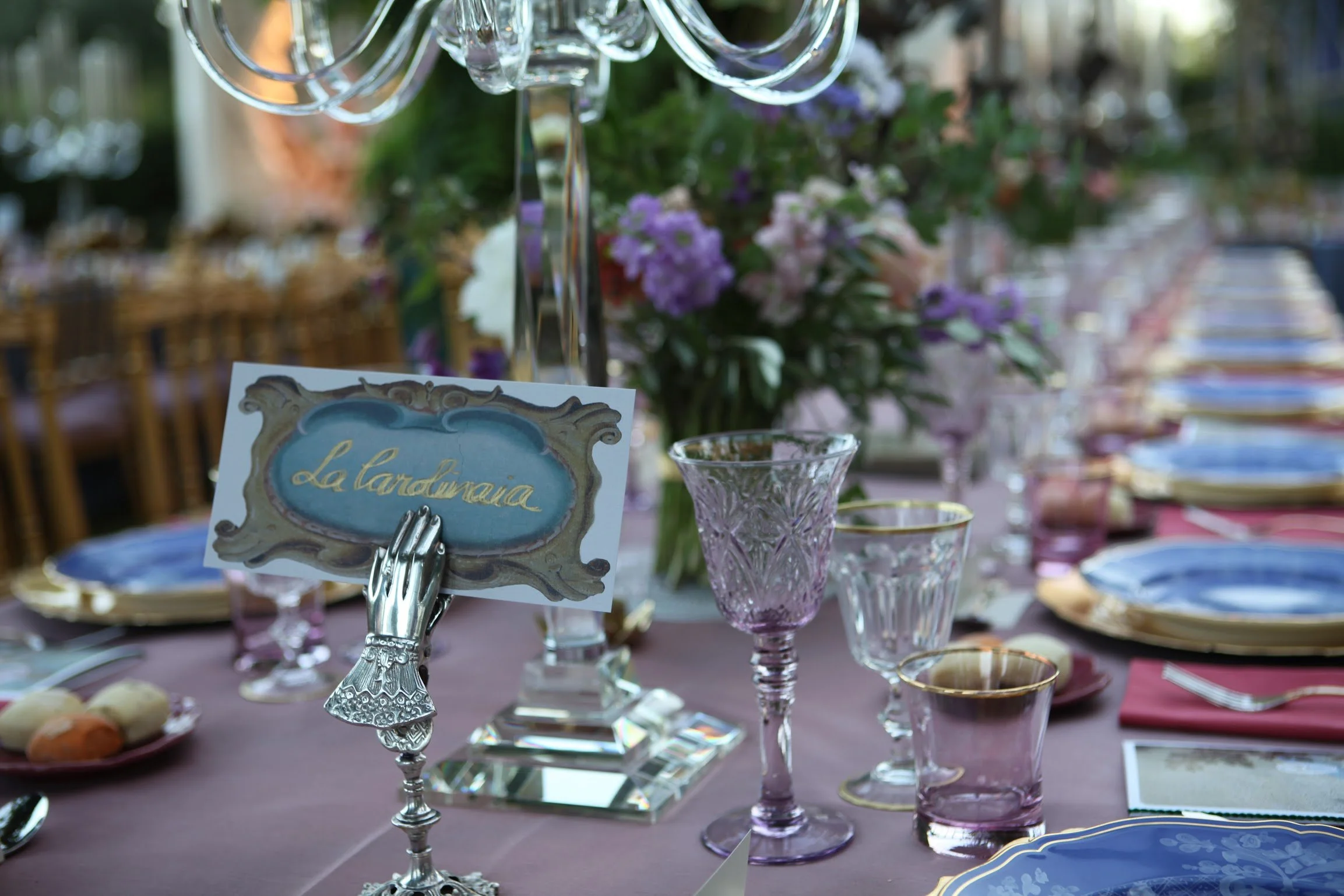 Close-up of a decorative silver spoon holding a card with the word "La tandorina" written in gold, surrounded by elegant purple glassware and floral arrangements on a table set for a formal event.