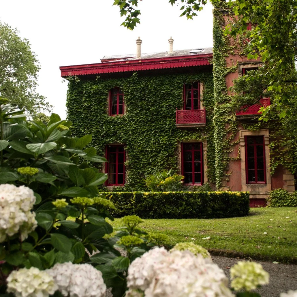 A two-story mansion, the Palacio de Arbaisenea,  covered in green ivy with red window frames and red balcony railings, surrounded by lush gardens and blooming flowers.