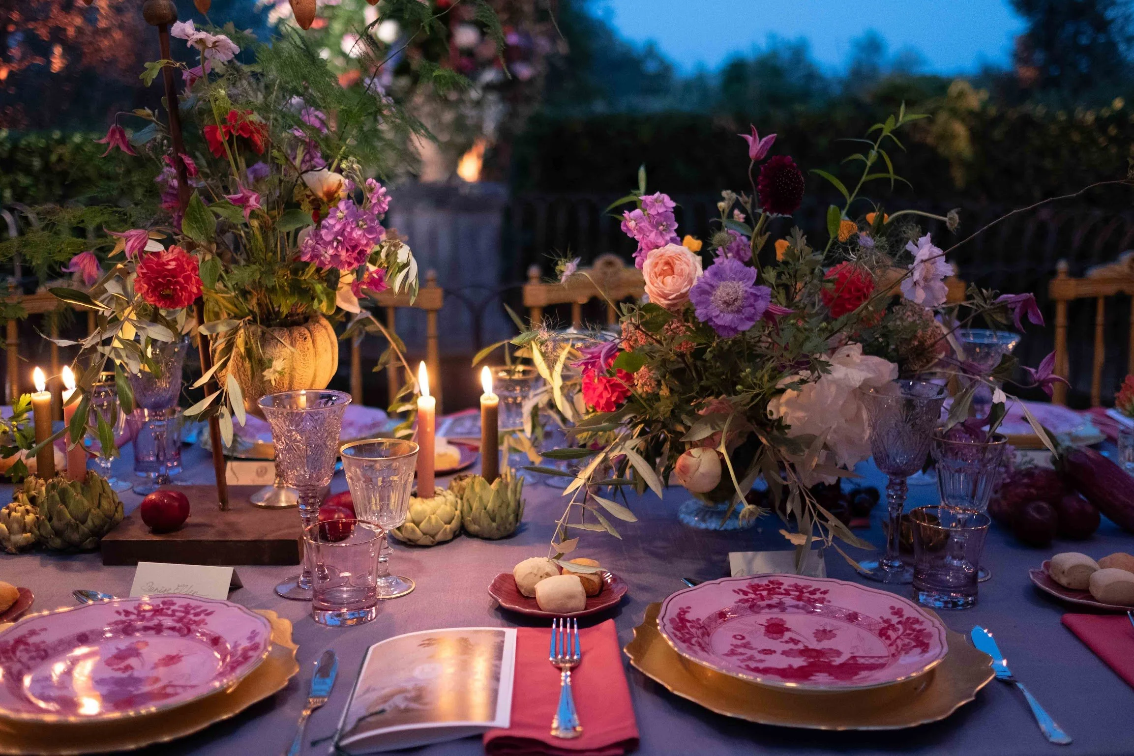 A beautifully decorated outdoor dining table at dusk, adorned with fresh flowers, candles, and elegant dinnerware, set for a special occasion or celebration.