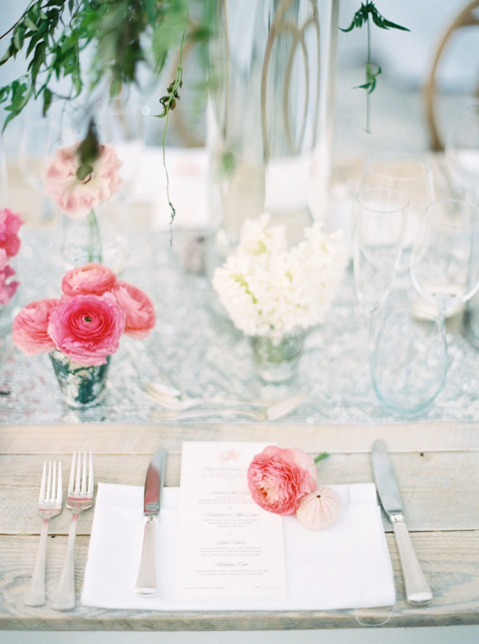 Table setting with pink and white flowers, glassware, and utensils, with a menu or program at the center.