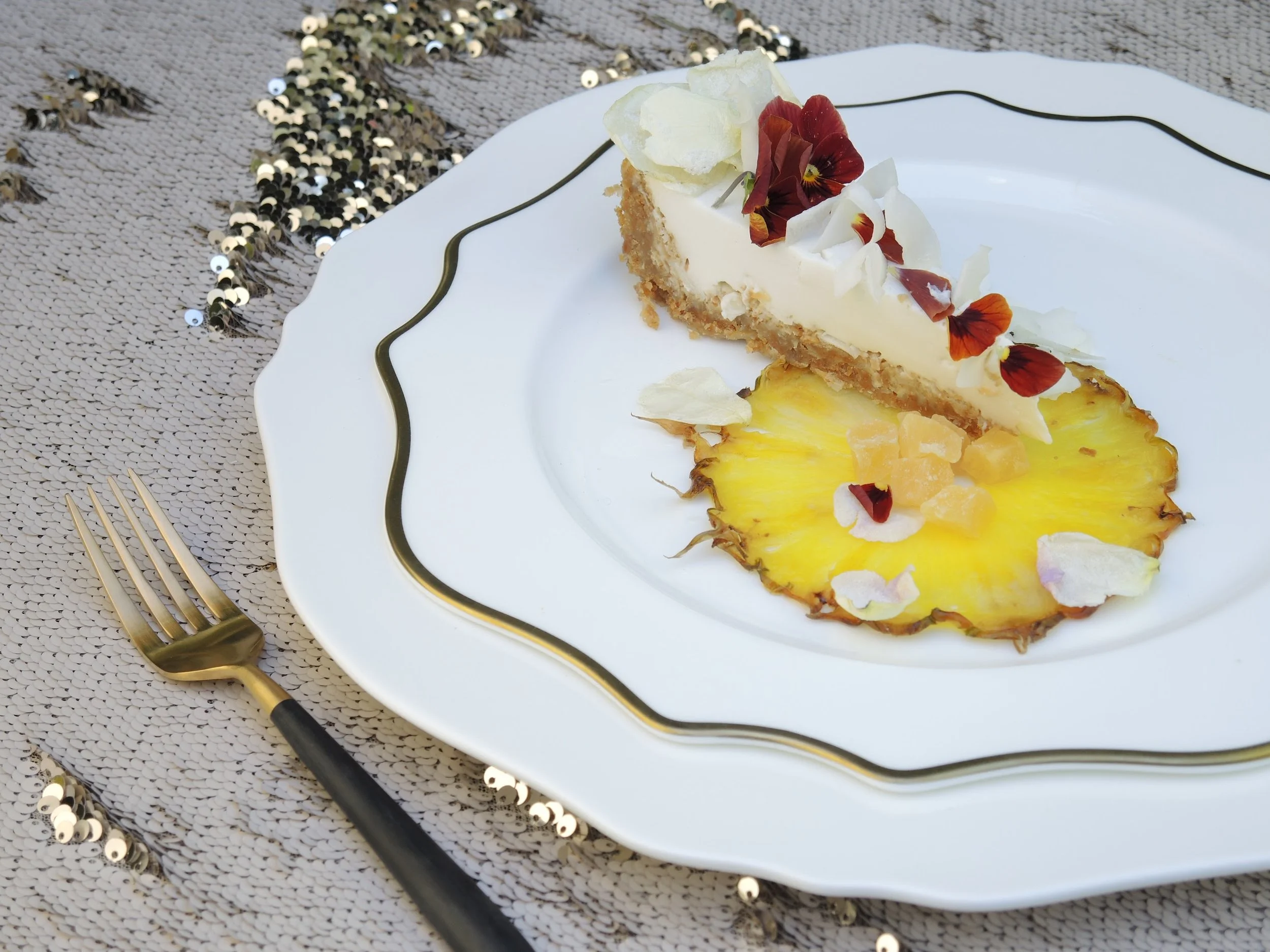 A slice of cheesecake topped with edible flowers on a white plate with gold trim, placed on a textured cream-colored tablecloth decorated with gold and black sequins, with a gold fork to the left.