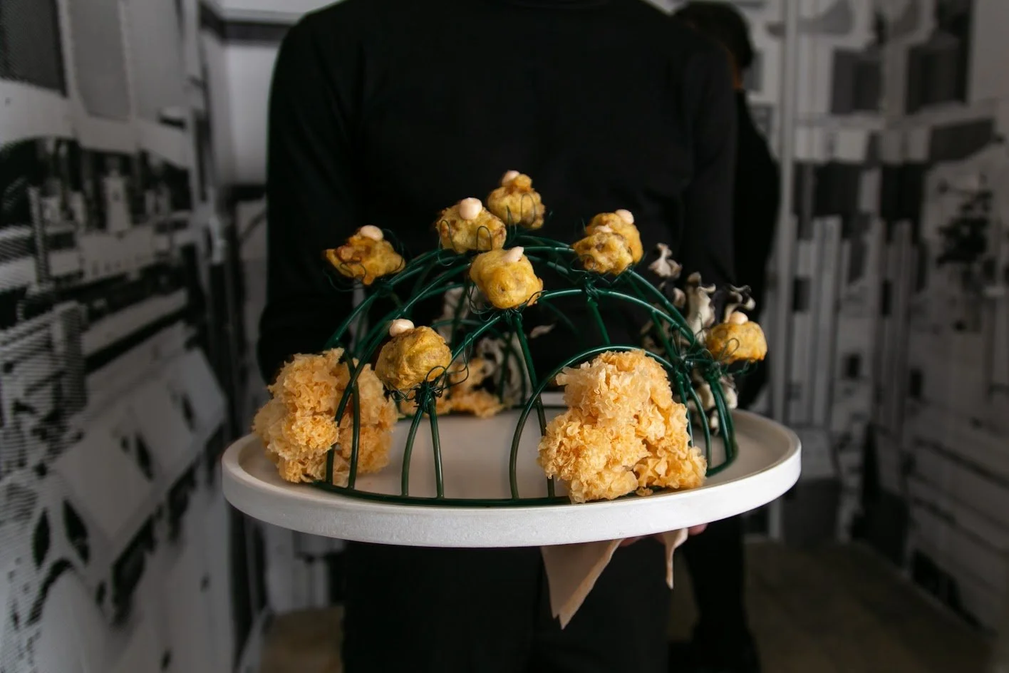 A person in black attire holding a white display tray with a decorative dome-like structure made of green wires adorned with small, yellow, flowering-looking elements and larger, fluffy yellow pom-pom flowers, set against a black and white patterned 