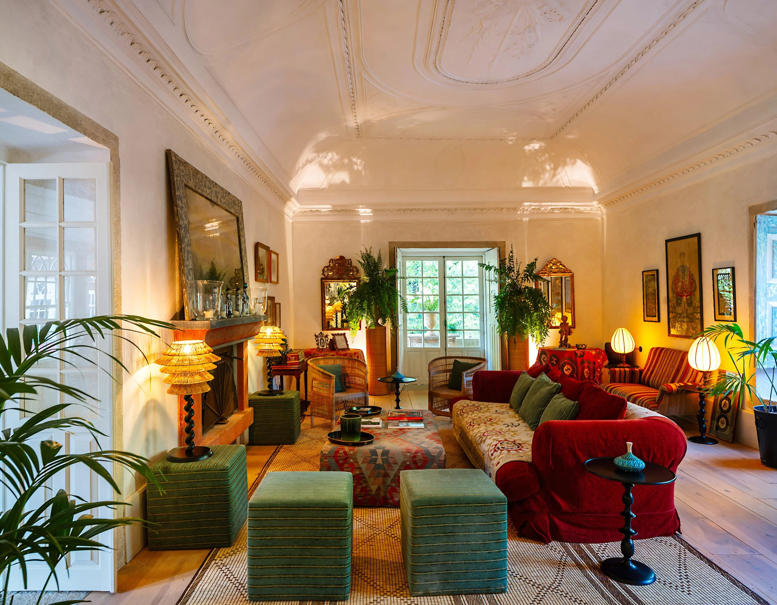 A cozy living room  at the Convento de Cabanas. with a high ornate ceiling, large windows, green and red upholstered furniture, potted plants, and various artwork on the walls.
