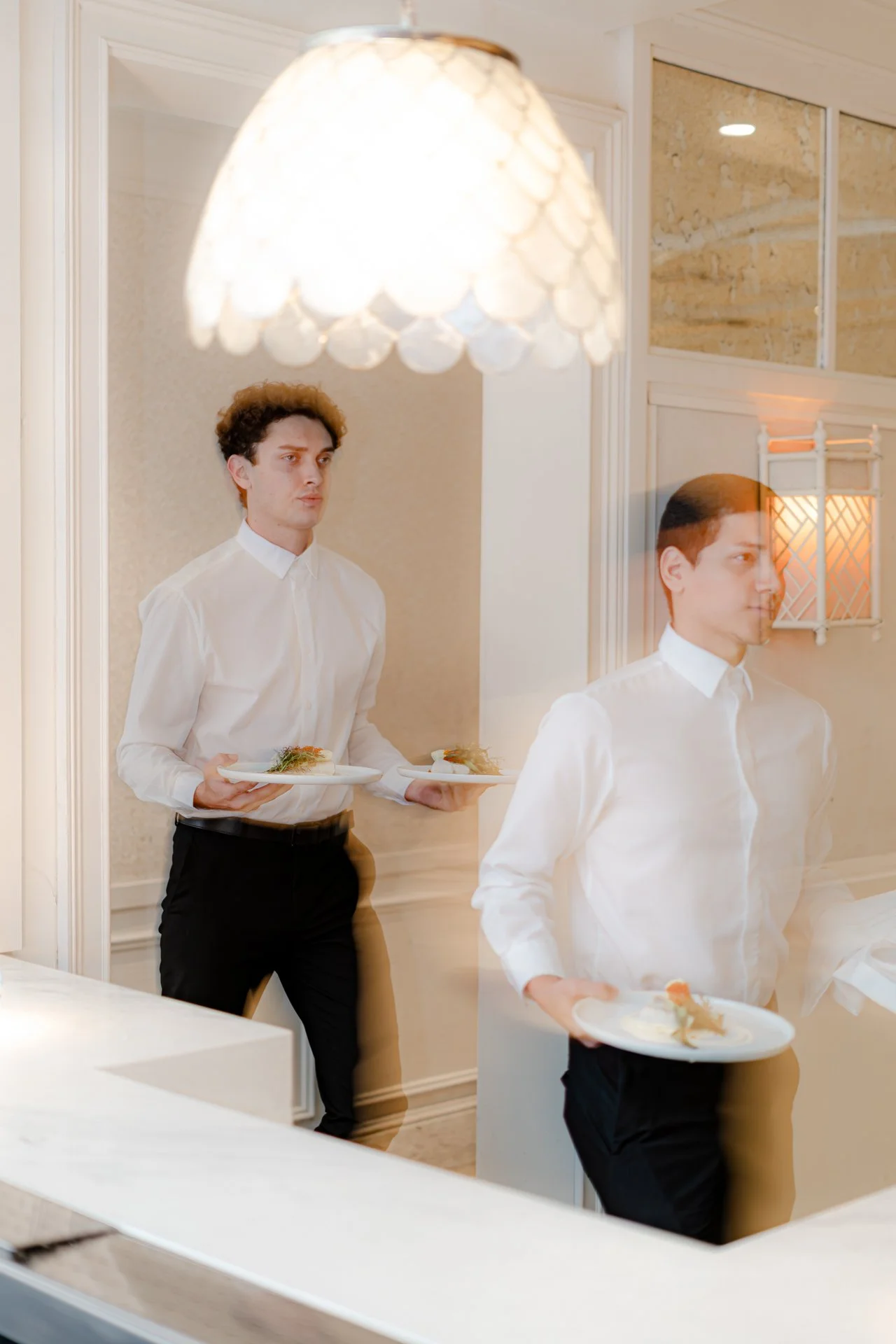Two young male servers dressed in white shirts and black trousers are reflected in a mirror, each holding a plate of food in a well-lit, elegant room with beige walls, white trim, and decorative mirrors.
