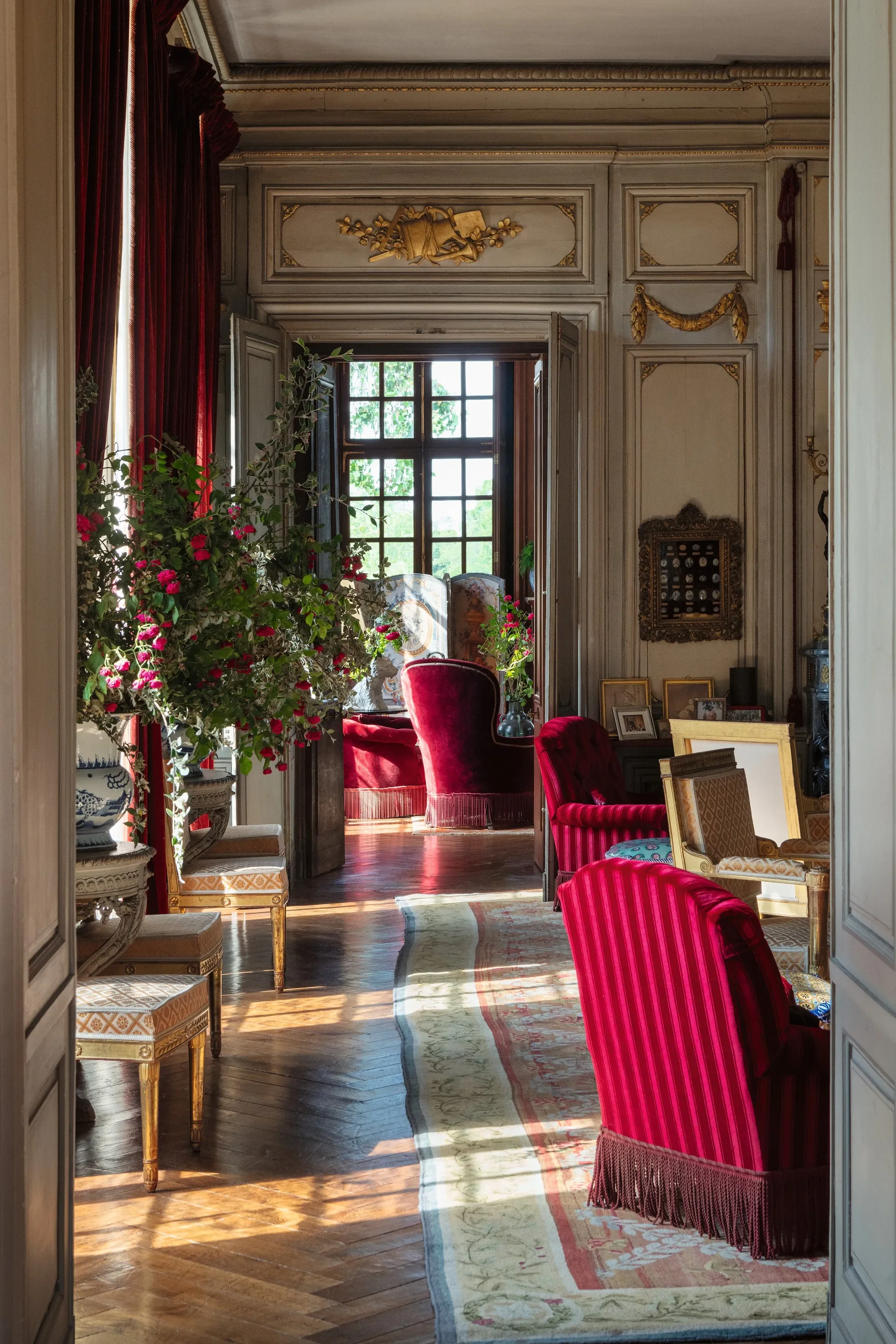 Elegant vintage living room with red velvet chairs, framed photos on a table, large window with views of greenery, and decorative gold accents on the walls and ceiling. The château is privately held but can host exclusive luxury events.