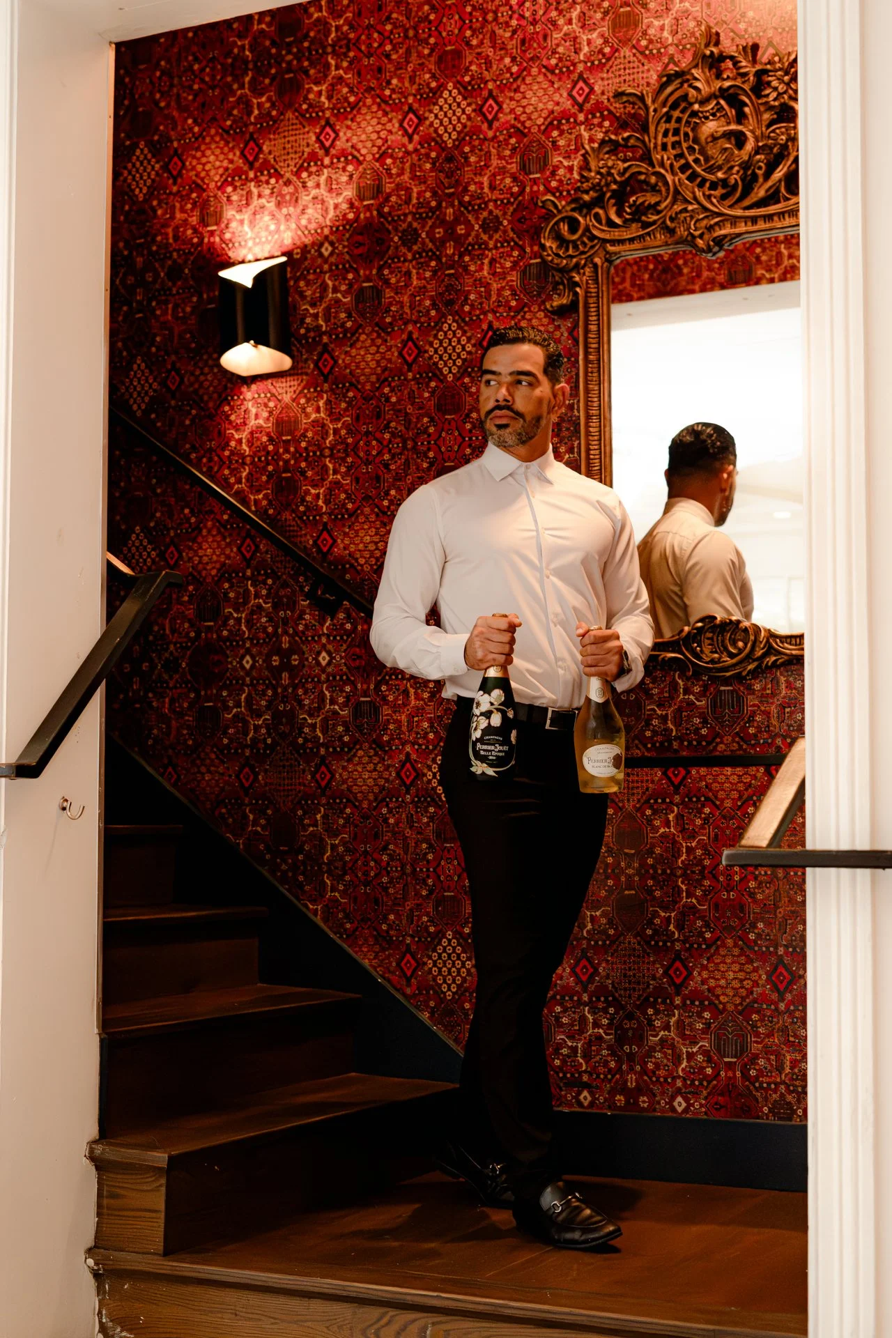 A man in formal attire holding two bottles of champagne standing on a staircase with a red patterned wall and ornate gold mirror in the background.