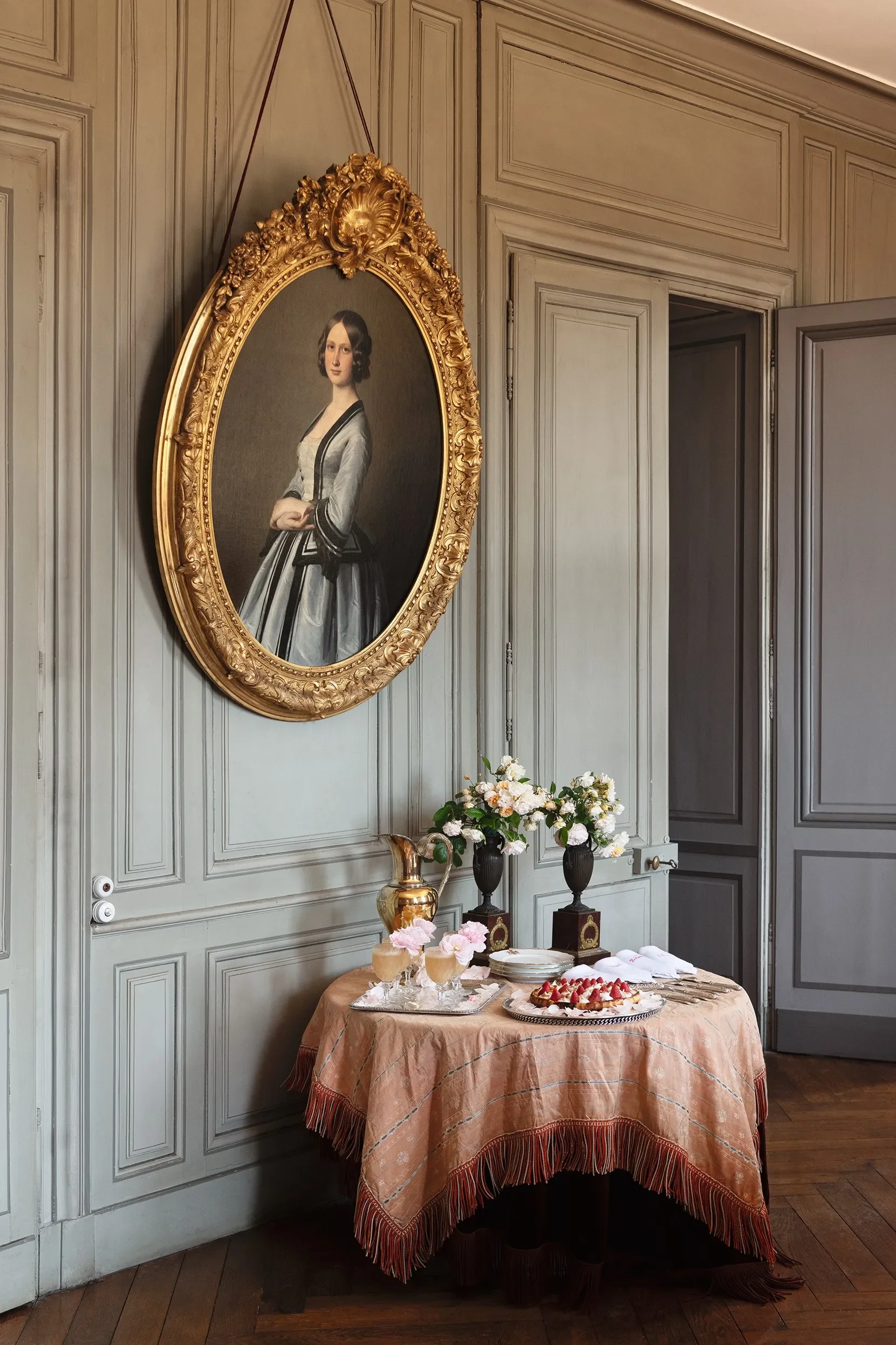 A room with gray paneled walls and an oval portrait of a woman in a black and white dress hanging on the wall. Below the portrait, a round table with a peach-colored cloth holds wine glasses with pink drinks, a cake with strawberries, flowers, and decorative vases.