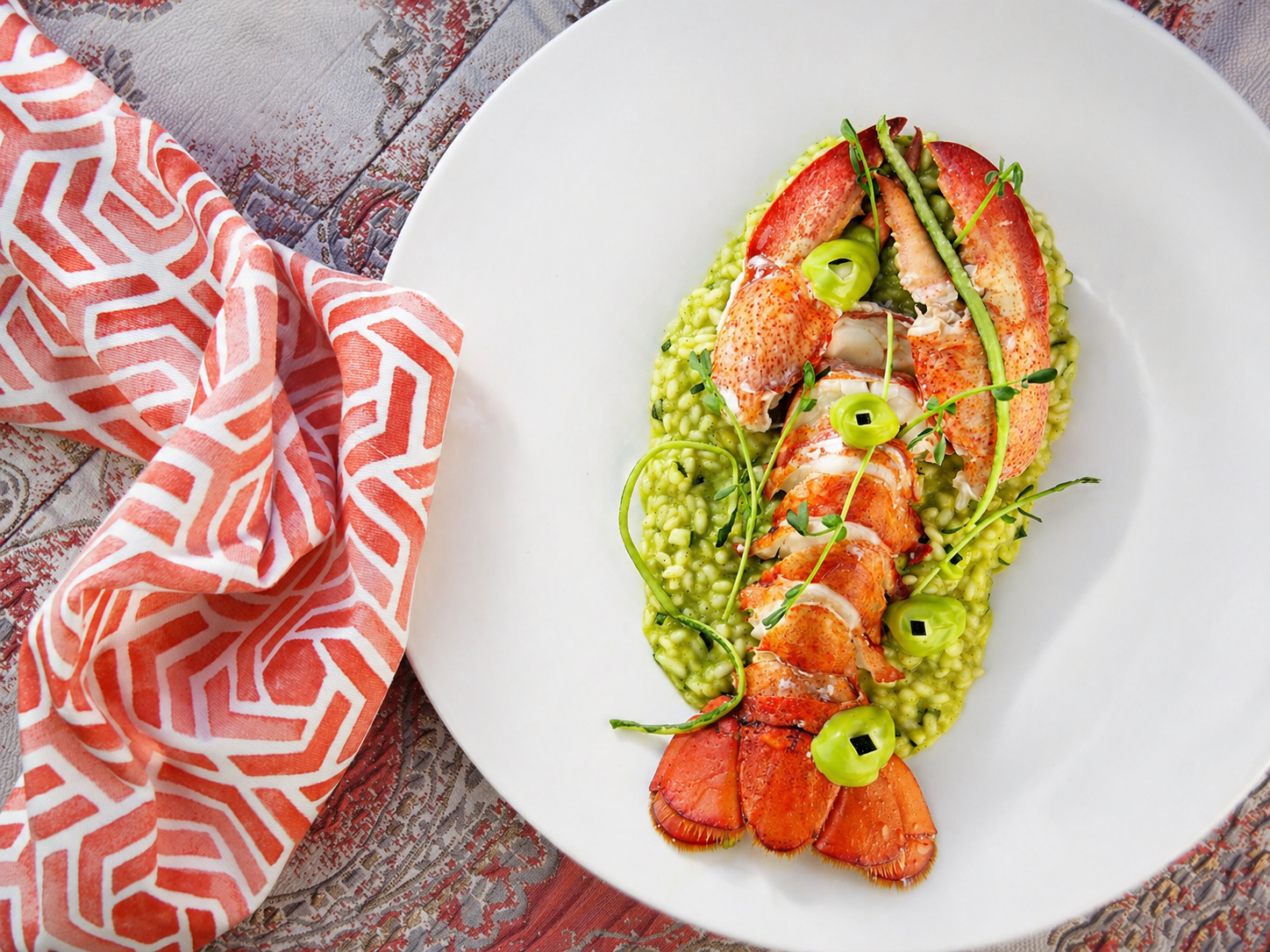 Lobster dish with green risotto garnished with microgreens and green onions on a white plate, placed on a textured surface with a red and white patterned cloth nearby.