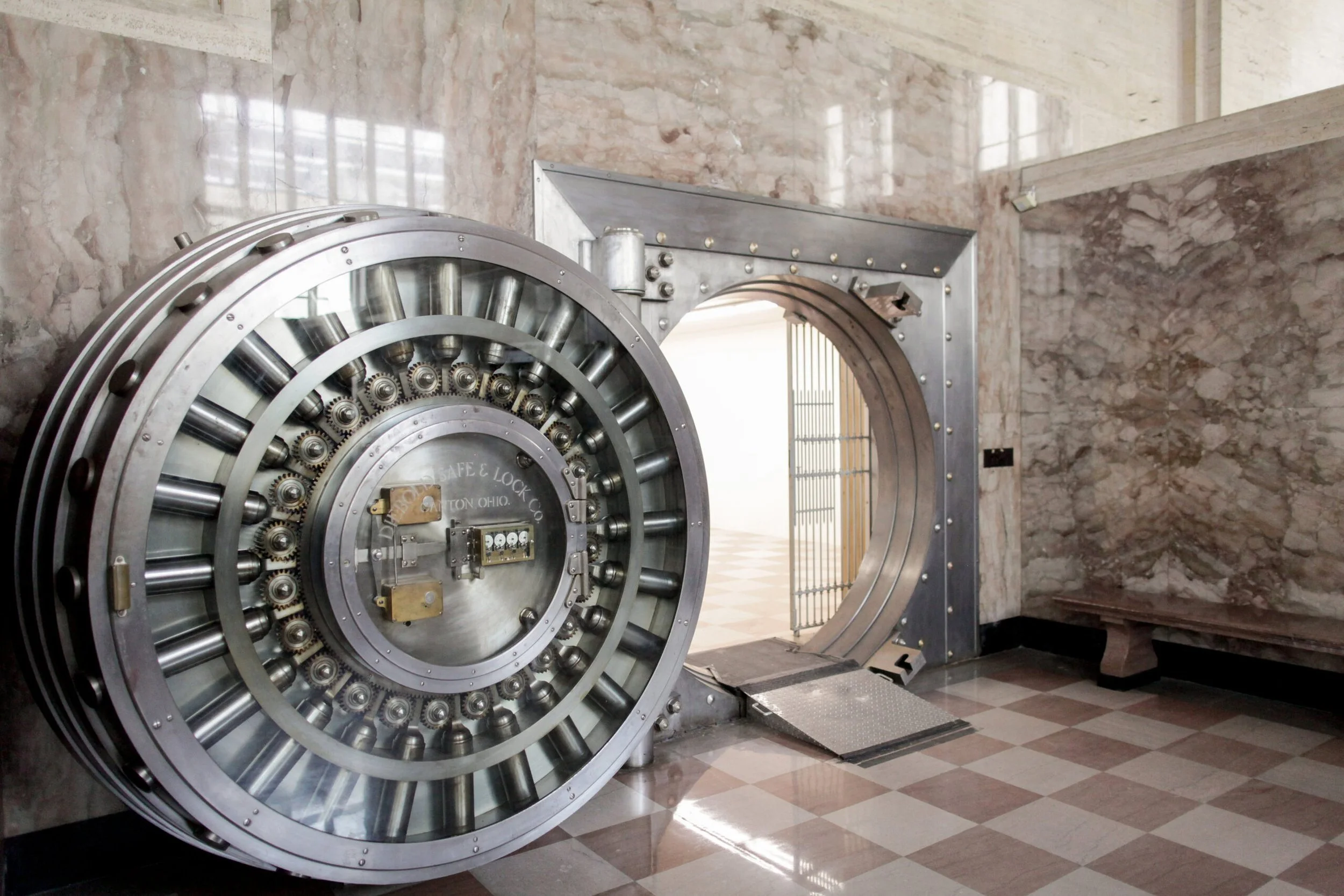 A large, secured, industrial-style vault door with a combination lock and gear mechanisms, situated inside a stone-walled room with a checkered tile floor and an open doorway leading to another room.