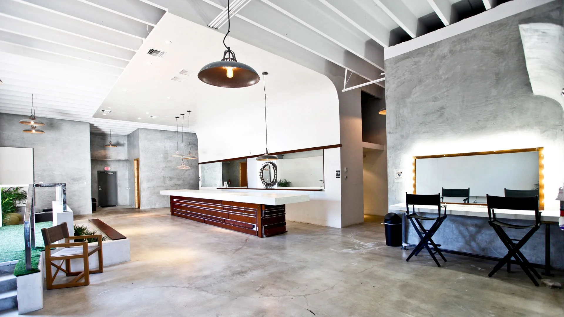 Modern interior space with concrete floors, white walls, pendant lighting, a reception desk, seating area with black chairs, a large mirror with lights, and decorative plants.