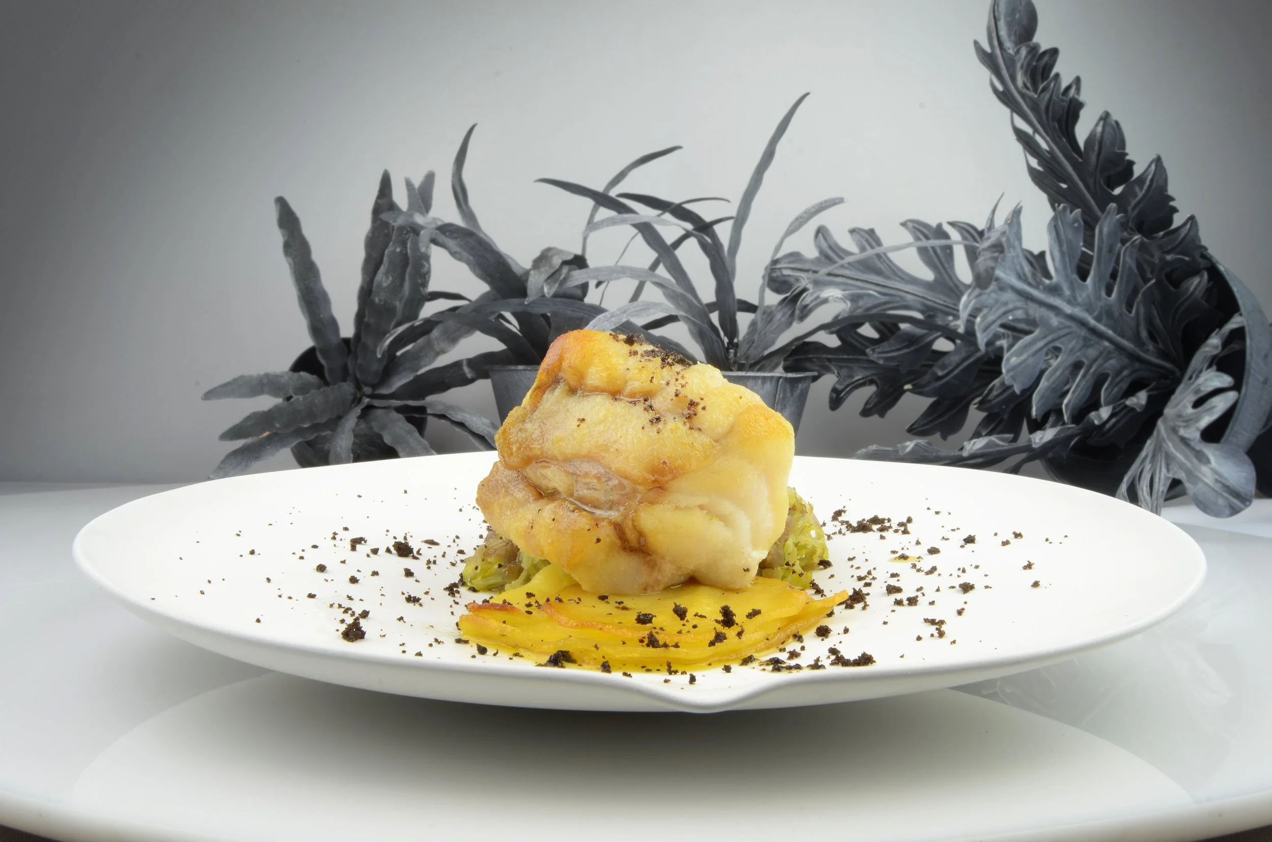 A plated dish featuring a piece of cooked chicken or fish on a bed of mashed potatoes, garnished with herbs and black crumbs, with black and gray tropical leaves in the background.
