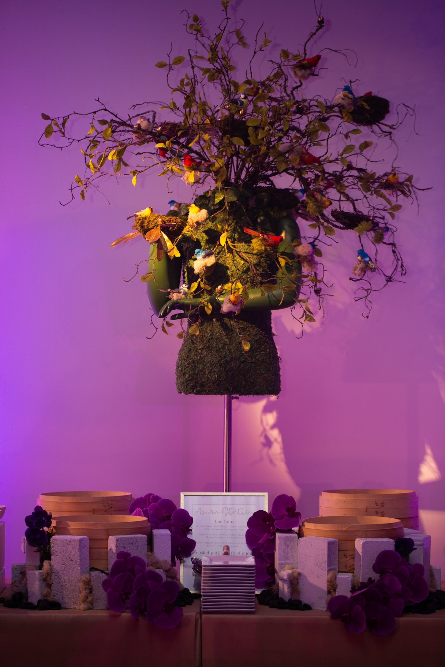 A decorative food display with a tree-shaped centerpiece adorned with small plush birds and leaves, surrounded by purple flowers, wooden crates, and a sign with menu items, illuminated with purple lighting.