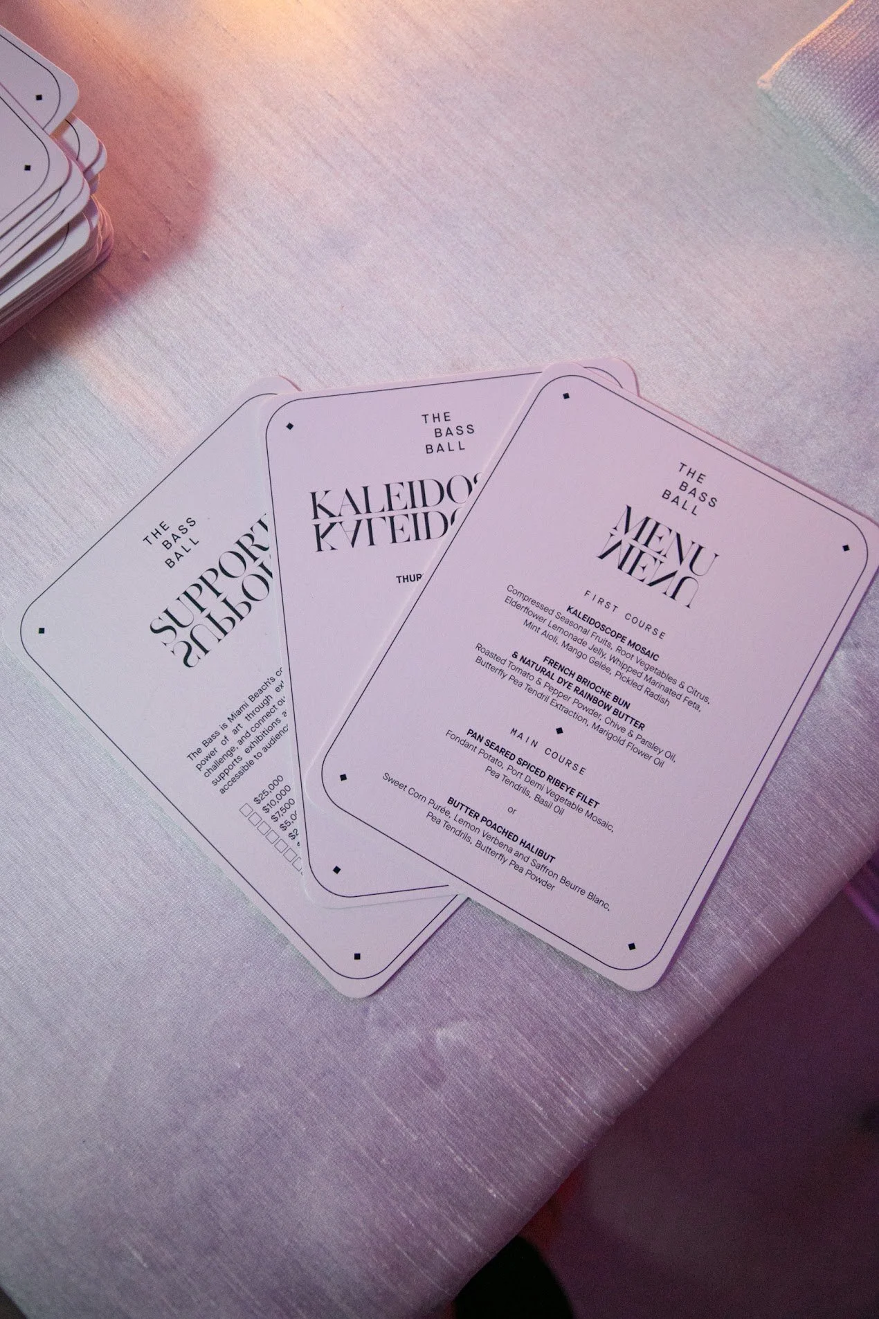 Three menus from The Bass Ball with black text on a white background, placed on a pink textured surface. The menus list courses and ingredients for a dining experience.