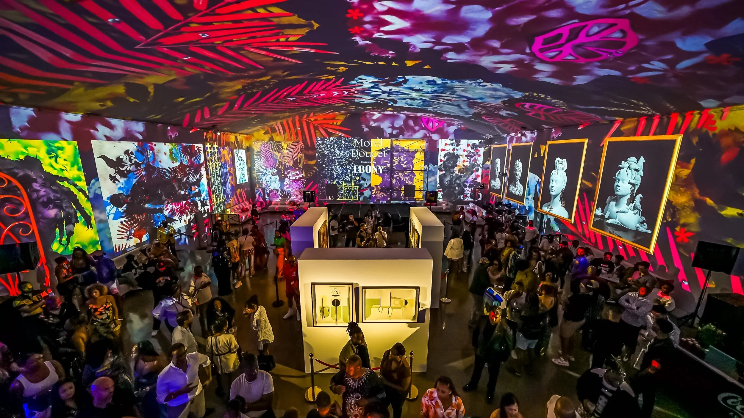 An indoor art exhibition with colorful wall and ceiling projections, featuring various artworks including digital prints and framed sculptures. Visitors are walking around and viewing the displays.