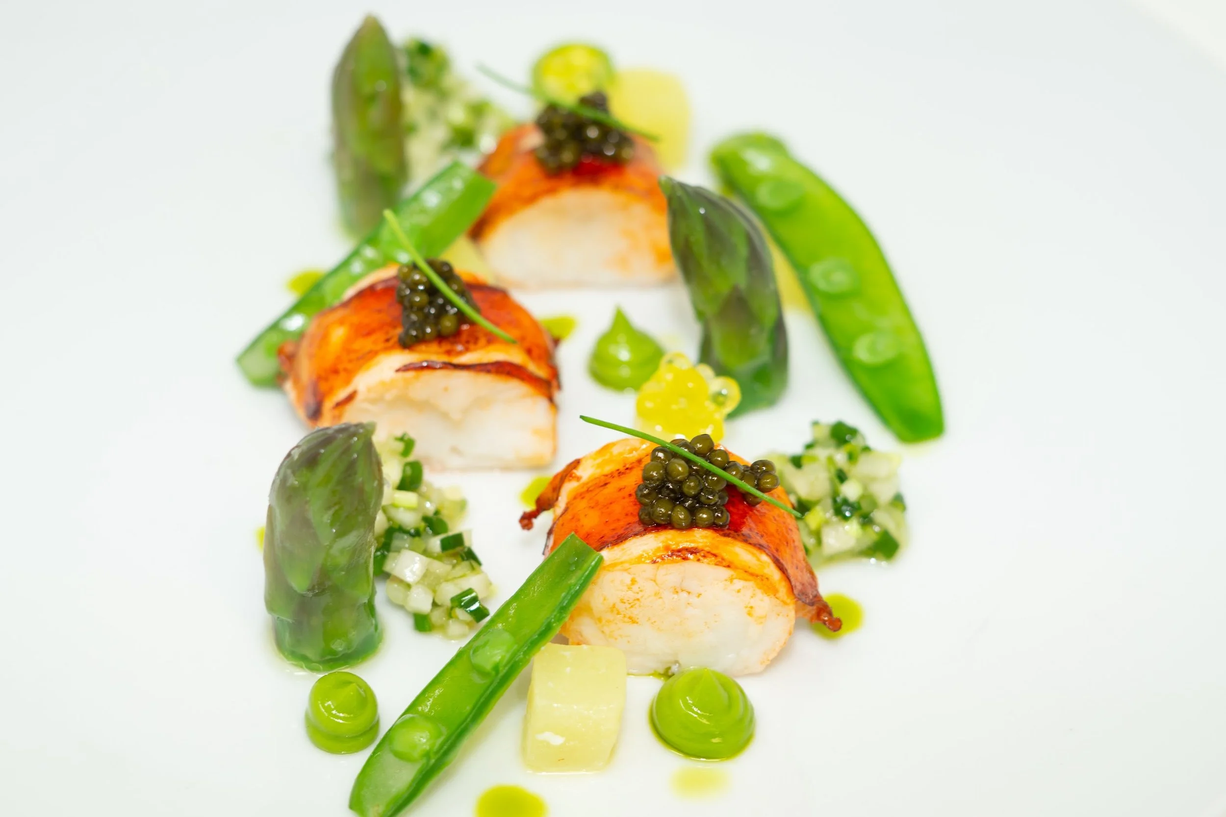 Gourmet dish featuring seared scallops wrapped in bacon, topped with black caviar, accompanied by green asparagus, diced vegetables, and drizzled with green sauce on a white plate.