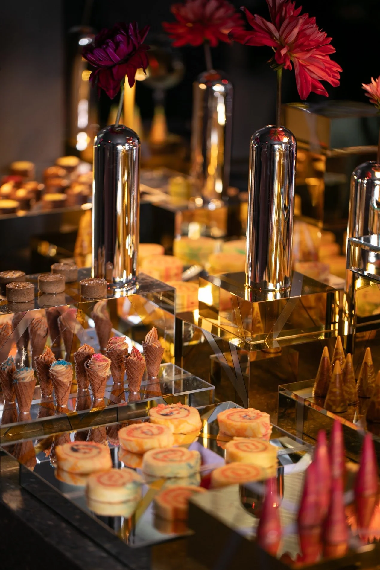 A display of assorted desserts including cupcakes, roll cakes, and ice cream cones on a tiered glass stand with metallic accents, decorated with vibrant red and purple flowers.