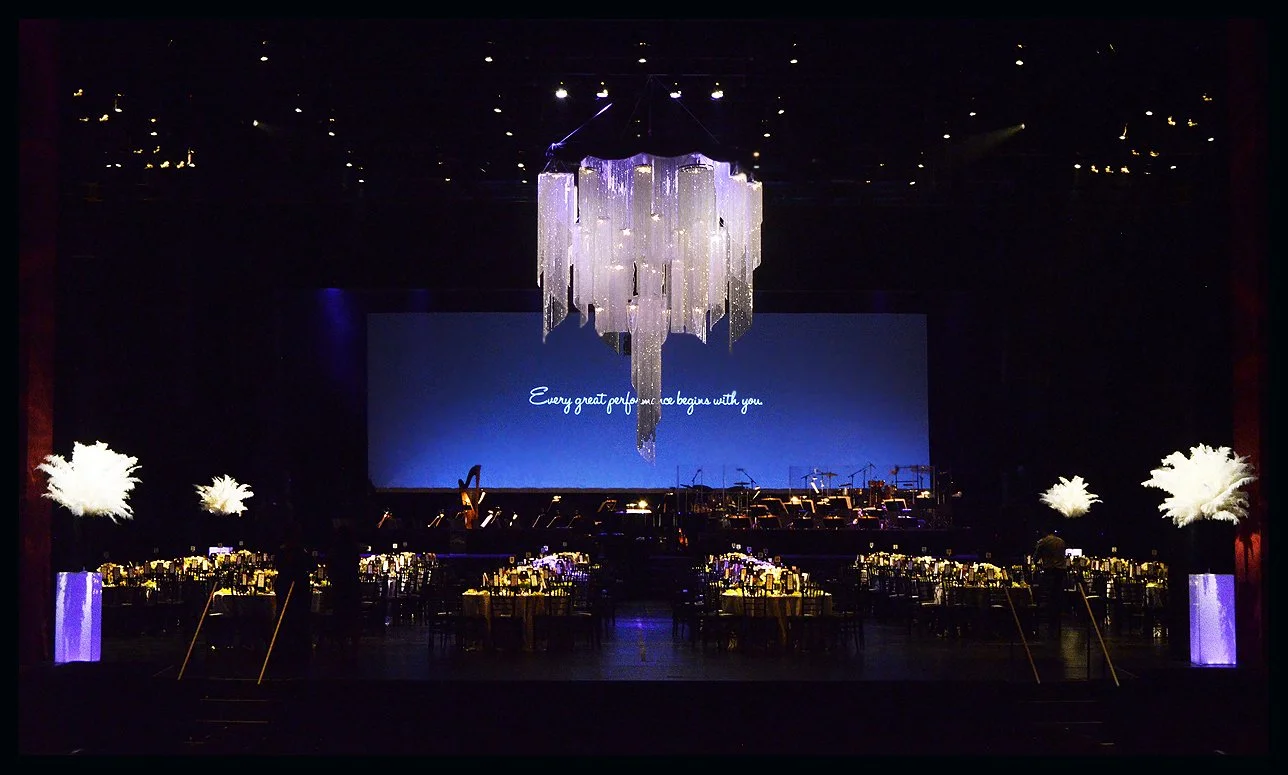 Elegant event hall with a large chandelier hanging from the ceiling, decorated tables with candle centers, and a large screen with the text "Every great performance begins with you."