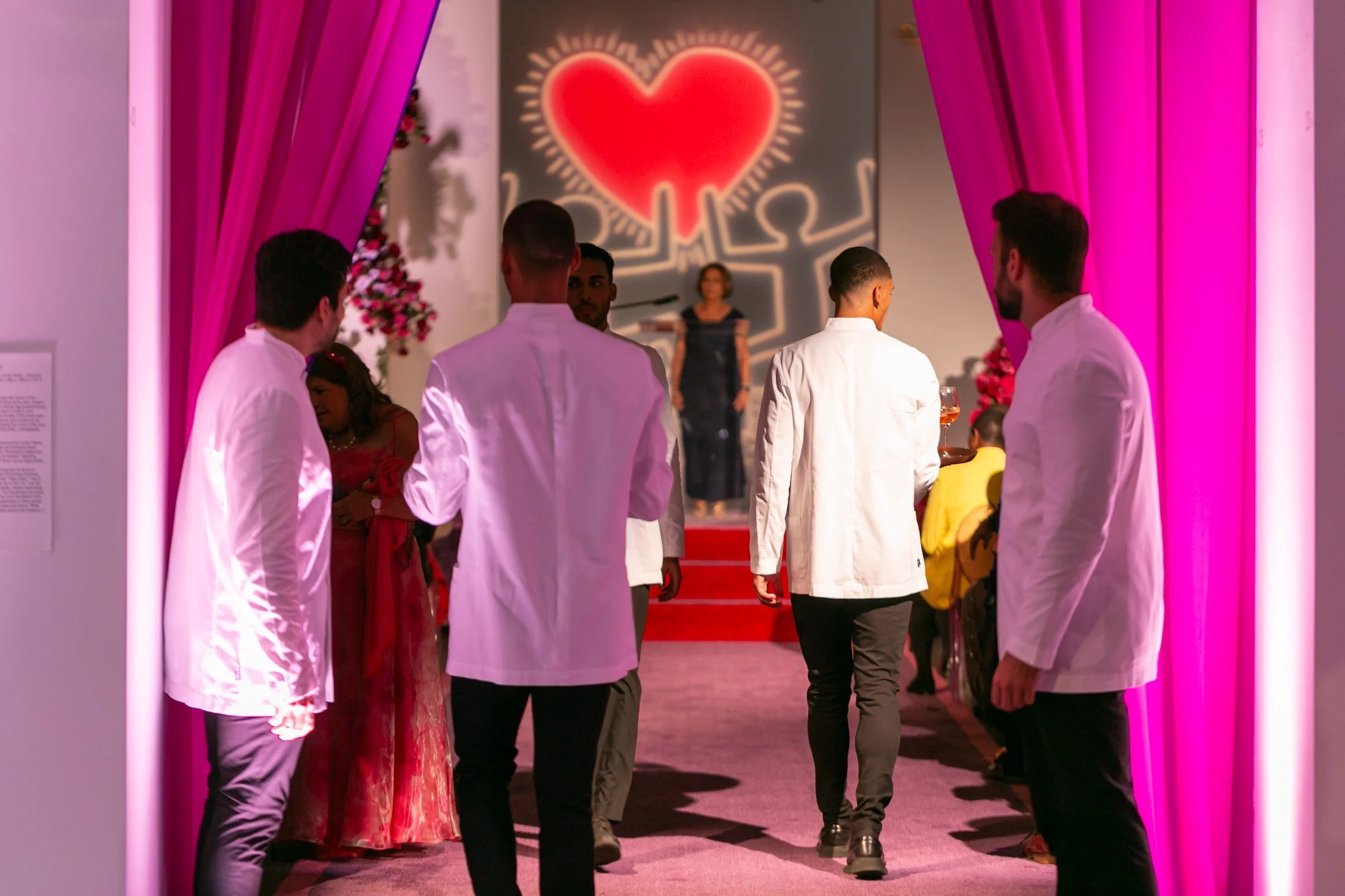 People in formal attire walking through entrance with pink curtains and a large red heart with a neon glow, background with pink and red decorations, possibly at a romantic or celebration event.
