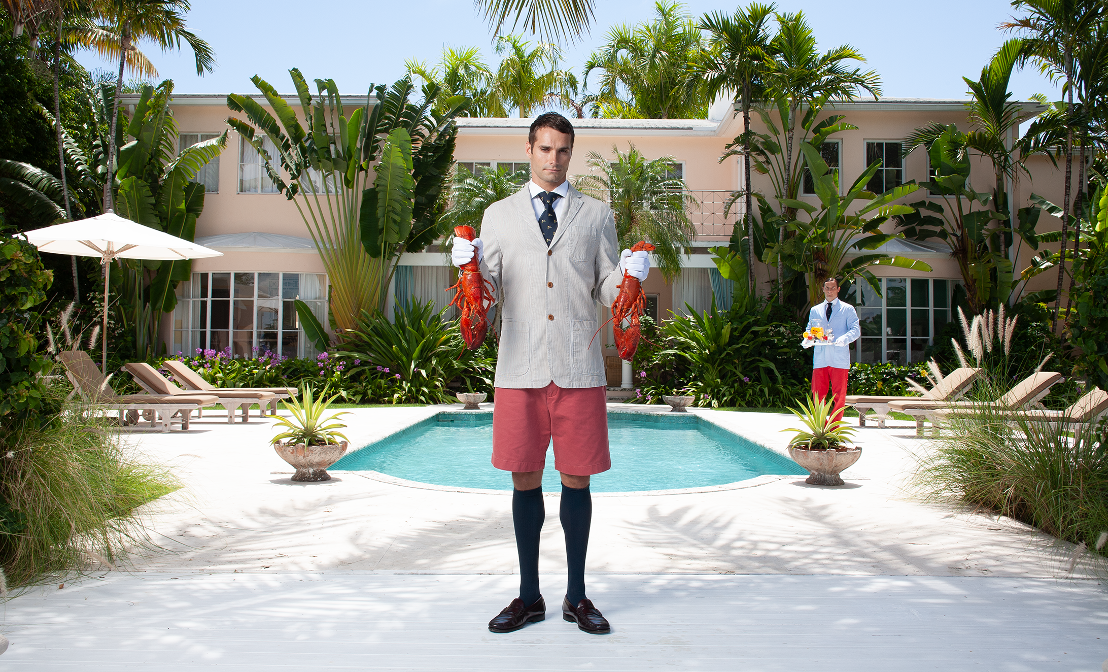 Man in gray jacket, red shorts, and tie holding two lobsters by a poolside with lush tropical plants and a pink building in the background.