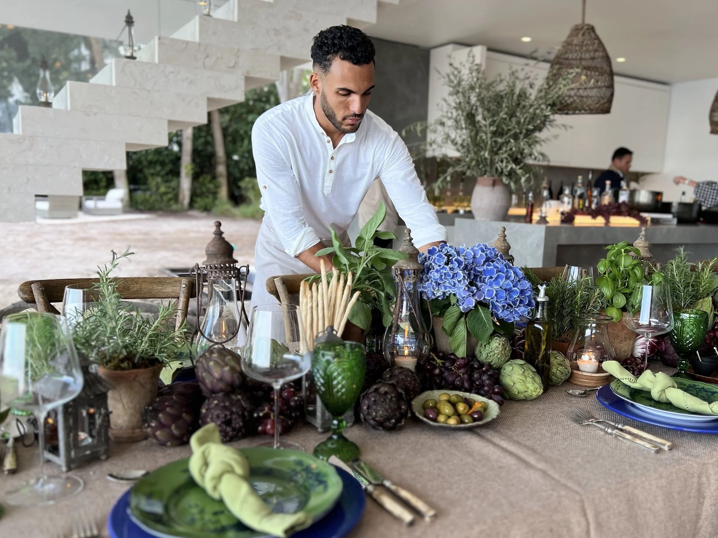 Man arranging a table of various plants, flowers, and food items at an indoor or outdoor dining event.